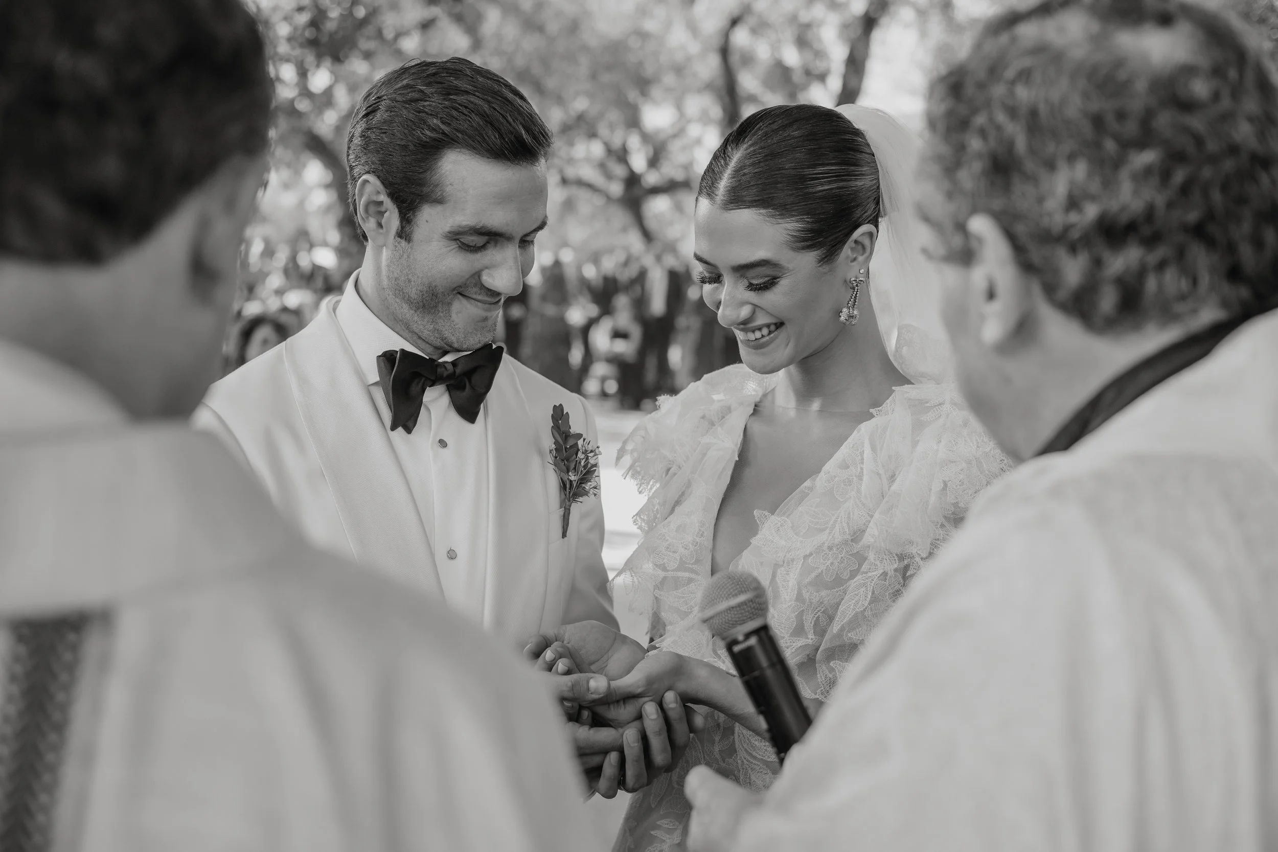 A black and white photo of a wedding ceremony outdoors with four people. The bride and groom are in the center, holding hands, smiling, with the officiant holding a microphone. Two other individuals are partially visible, facing the couple.