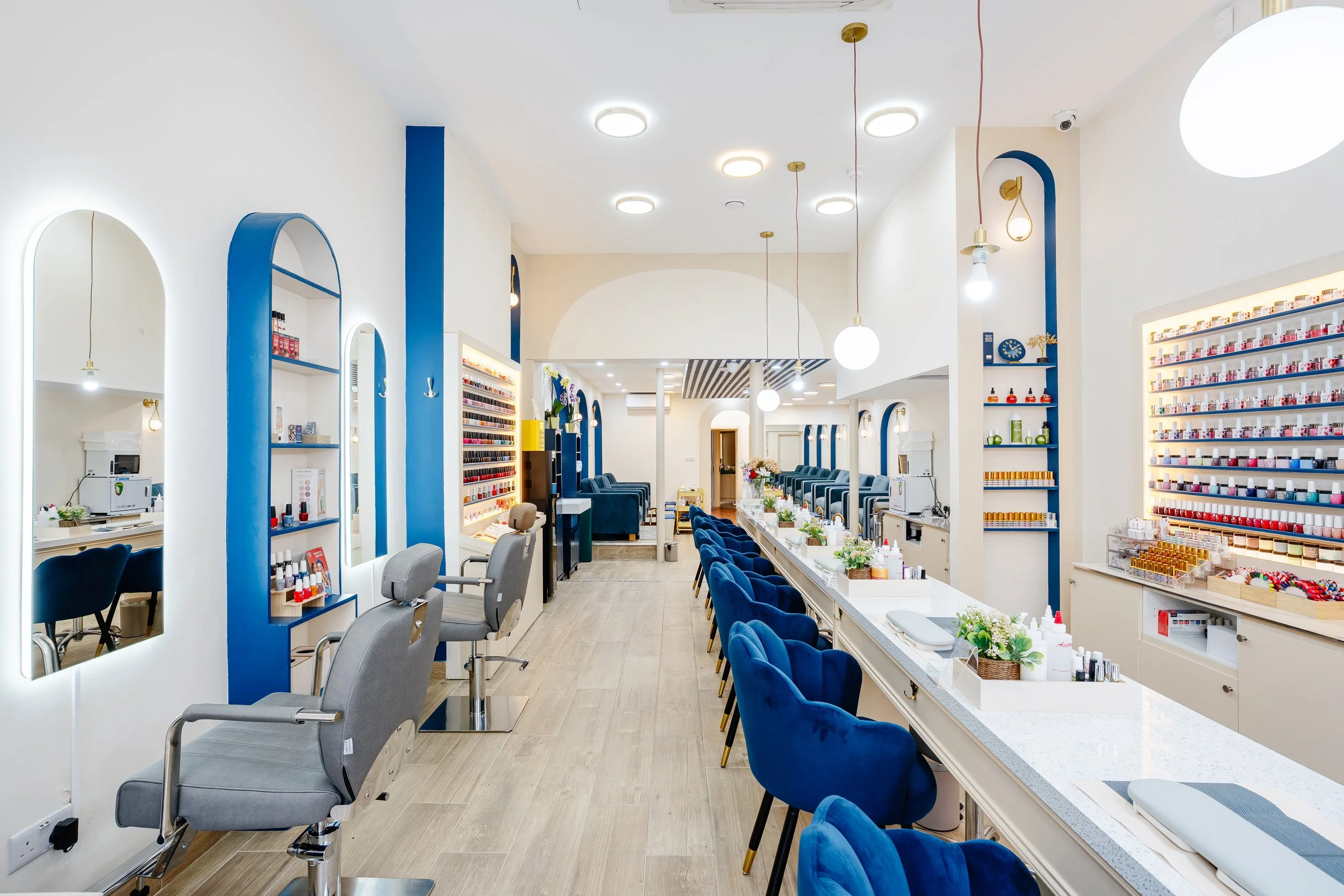 Interior of a nail salon with white walls, blue accents, and a long row of blue velvet chairs at manicure stations with nail polish racks on the walls.