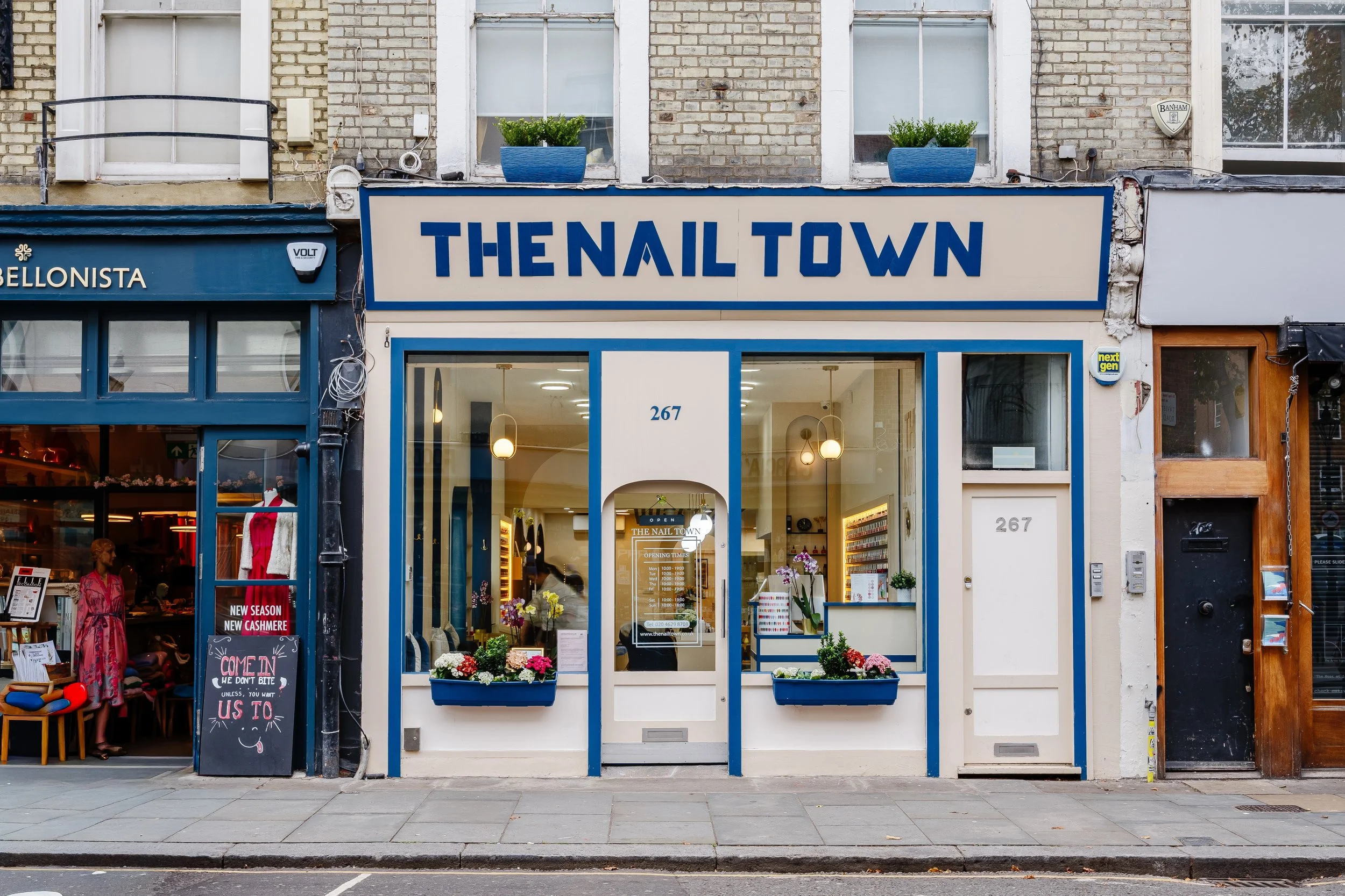 storefront with a sign reading 'The Nail Town', decorated with flowers, with an entrance and large windows, located at 267, with neighboring shops.