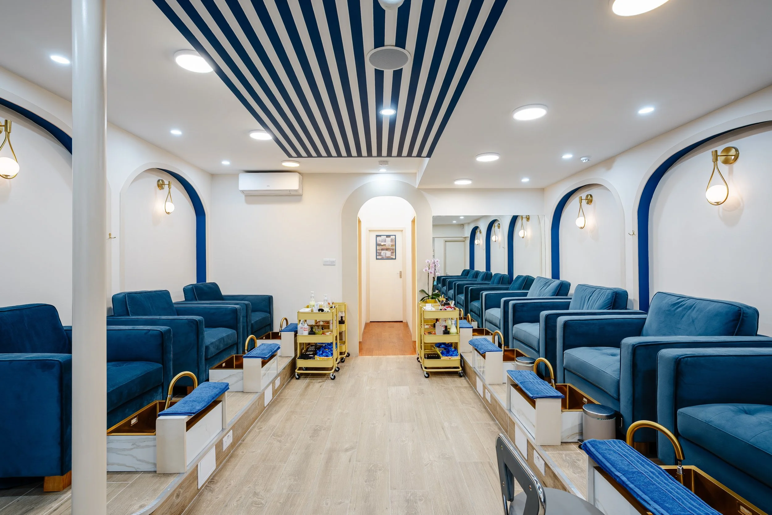 Room with multiple blue reclining chairs along the walls, foot baths, and small carts with towels and amenities, with a door at the end.