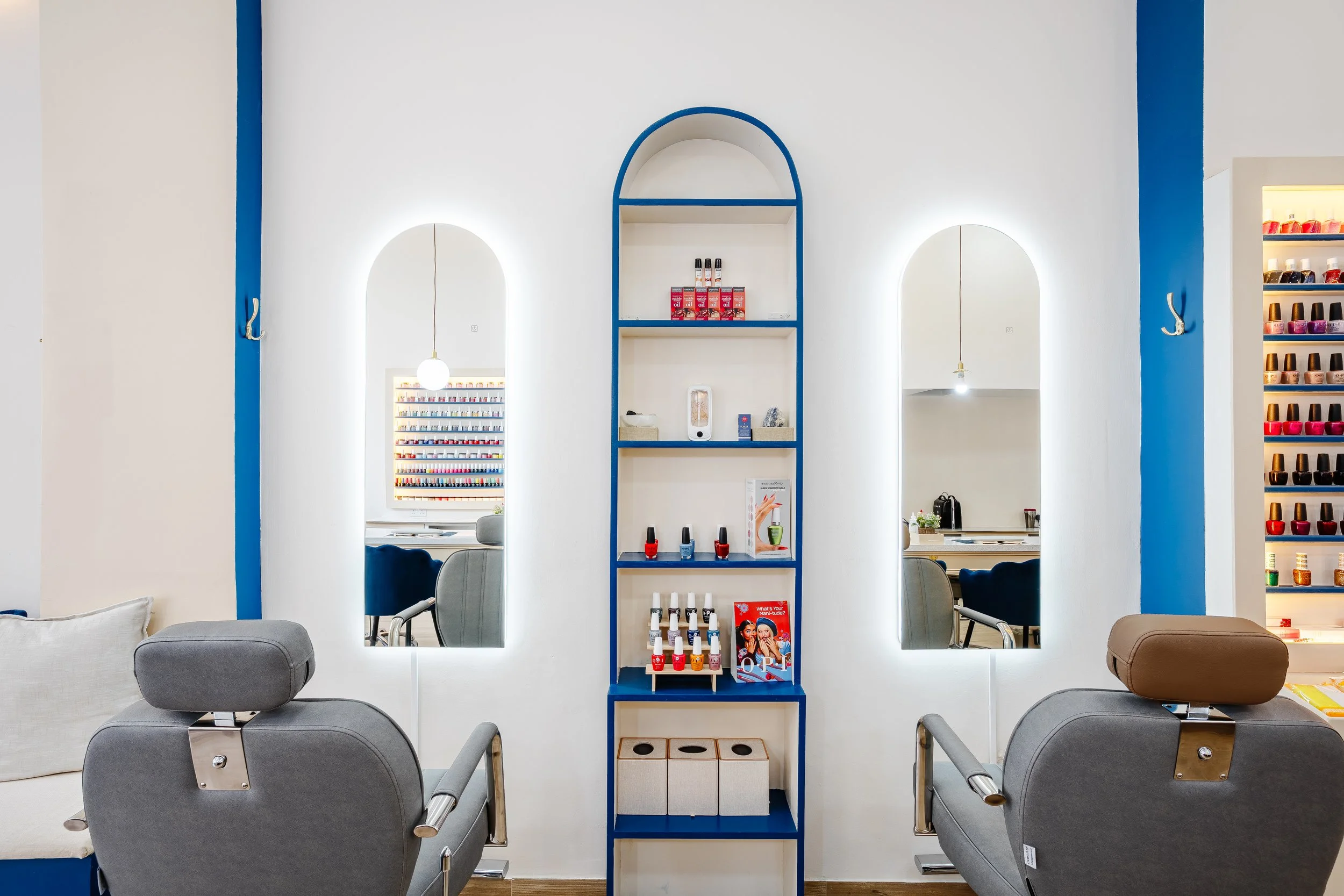 Salon with two grey chairs, a white pillow, and a blue and white wall with shelves and mirrors, nail polish displays, and salon equipment.