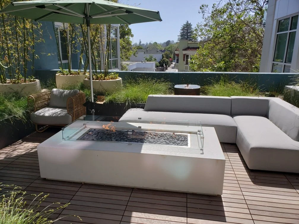Outdoor rooftop patio with a large L-shaped white sectional sofa, a glass-top fire pit table, a wicker armchair, and large potted plants with greenery, under a green umbrella, overlooking a neighborhood with trees and buildings.