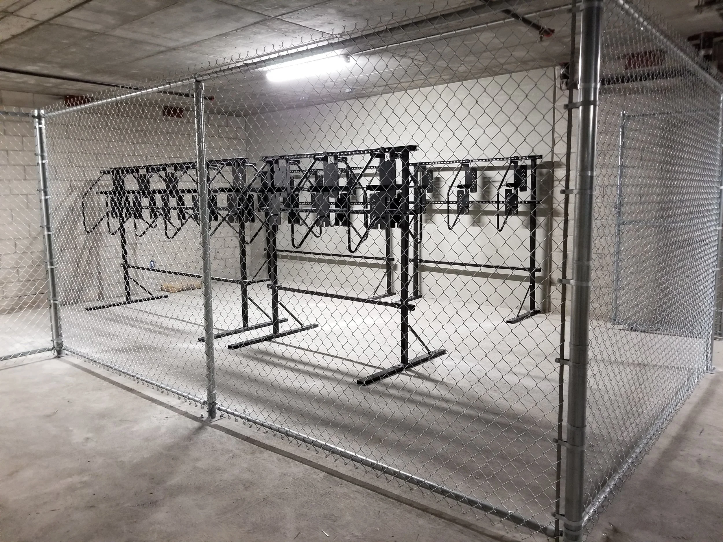 A fenced-off room with a chain-link fence and metal poles, containing multiple black bike racks with hangers, in a basement or storage area.