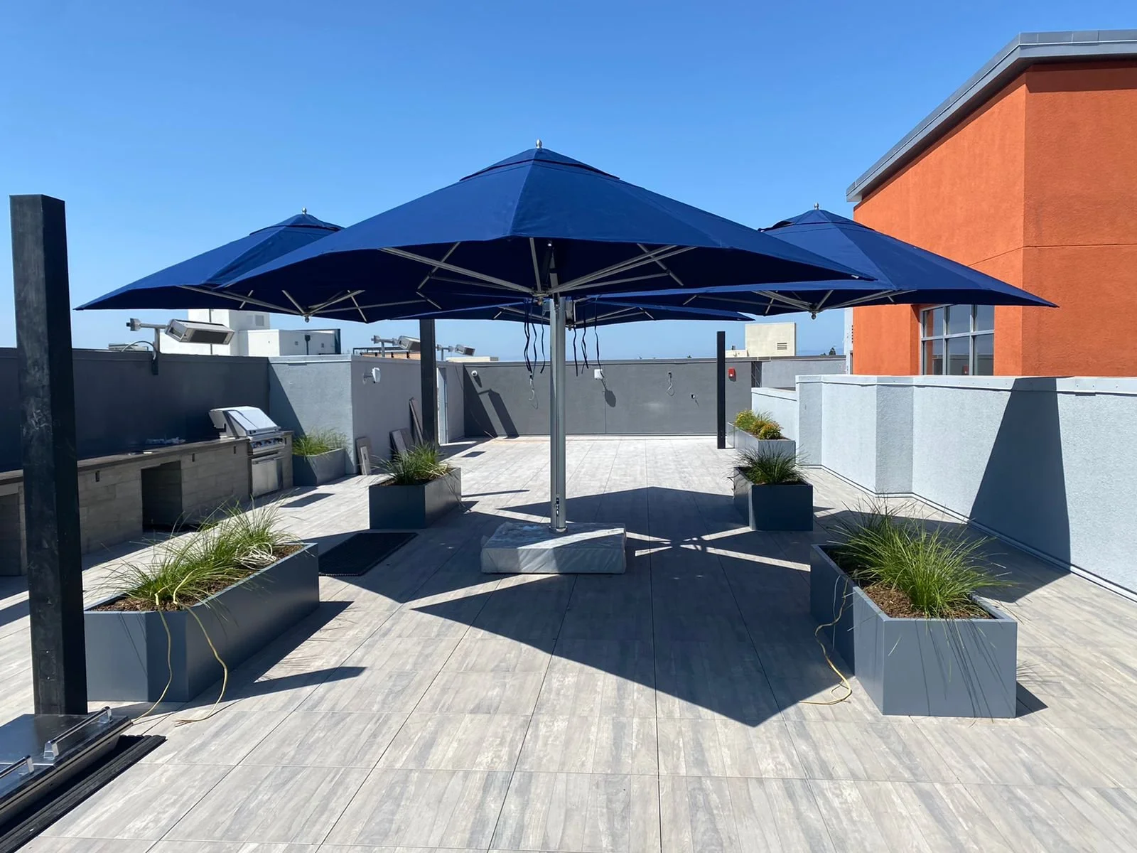 Outdoor rooftop patio with large blue umbrellas, potted modern planters with grass, built-in grill area, gray and orange walls, clear blue sky.