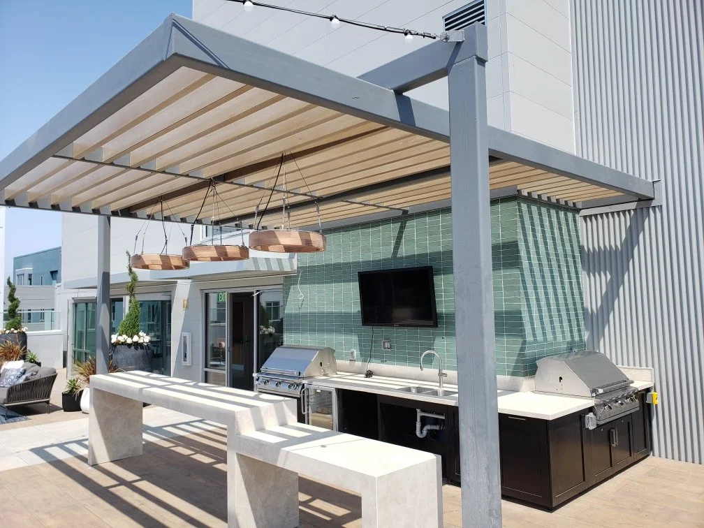 Outdoor kitchen with two grills, a green tiled wall, a wall-mounted TV, and a shaded pergola in a rooftop setting.