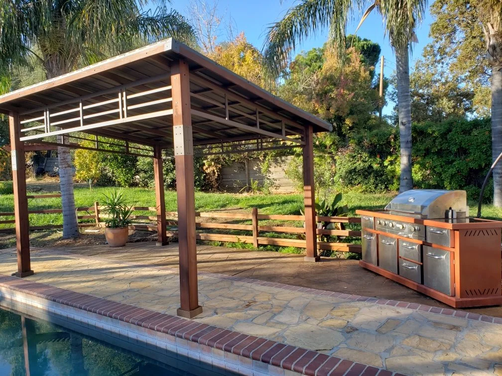 Backyard patio with a canopy shade structure, potted plant, built-in grill, and lush trees in the background.