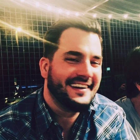 A smiling man with dark hair and a beard, wearing a blue plaid shirt.