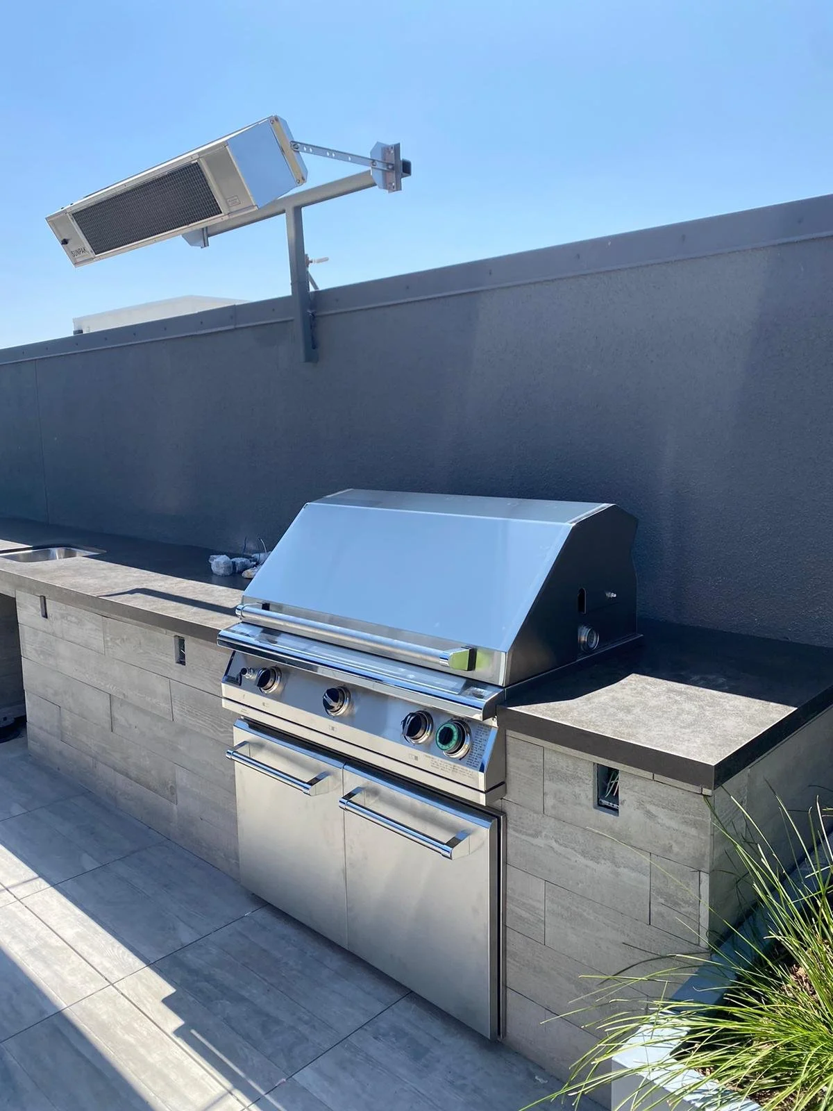 Stainless steel outdoor gas grill on a patio with a gray stone countertop, set against a dark gray wall, under clear blue sky.