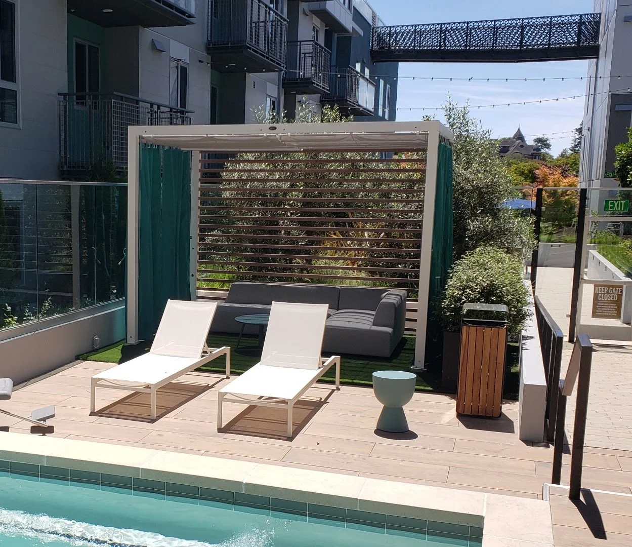 Poolside outdoor seating area with two white lounge chairs, a gray sofa under a shaded cabana, a small round light blue table, and a wooden trash bin, surrounded by a modern apartment building with balconies and a bridge structure overhead.