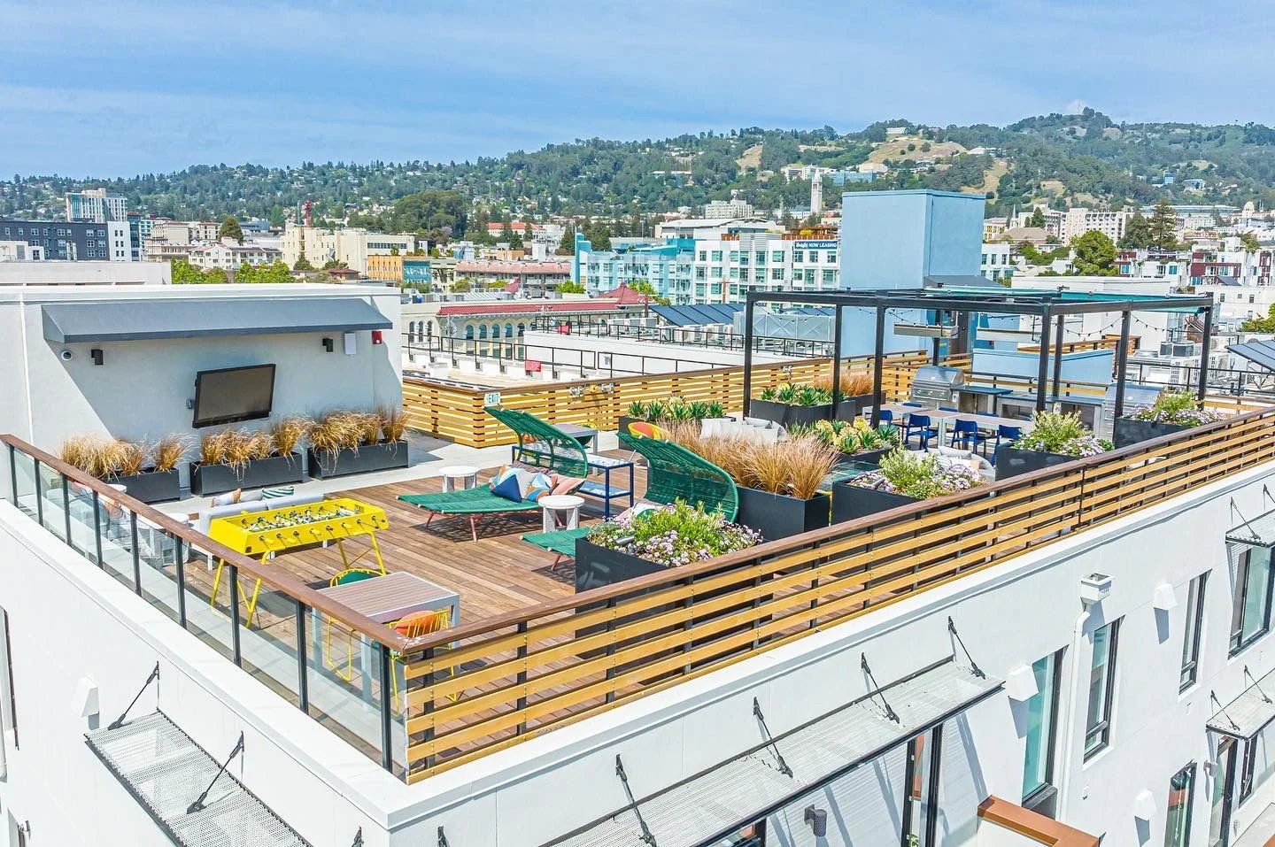 Rooftop with outdoor furniture, plants, lounge chairs, and a foosball table in an urban area under a blue sky.
