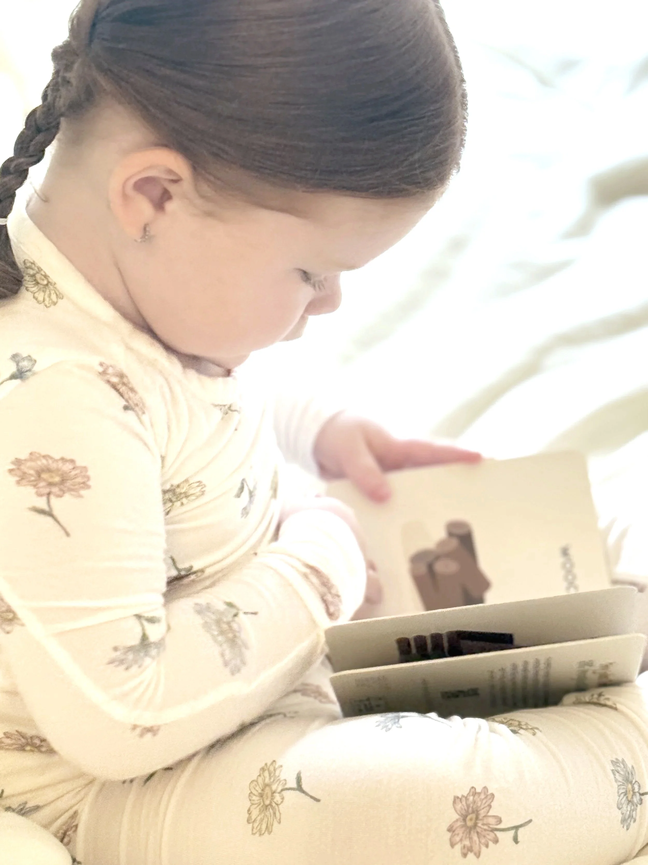 A young child with braided hair reading an illustrated board book from the Little Life Series by Sunshine & Poppies Press. Soft lighting and neutral tones create a calm, story-time moment for early readers ages 0–3.