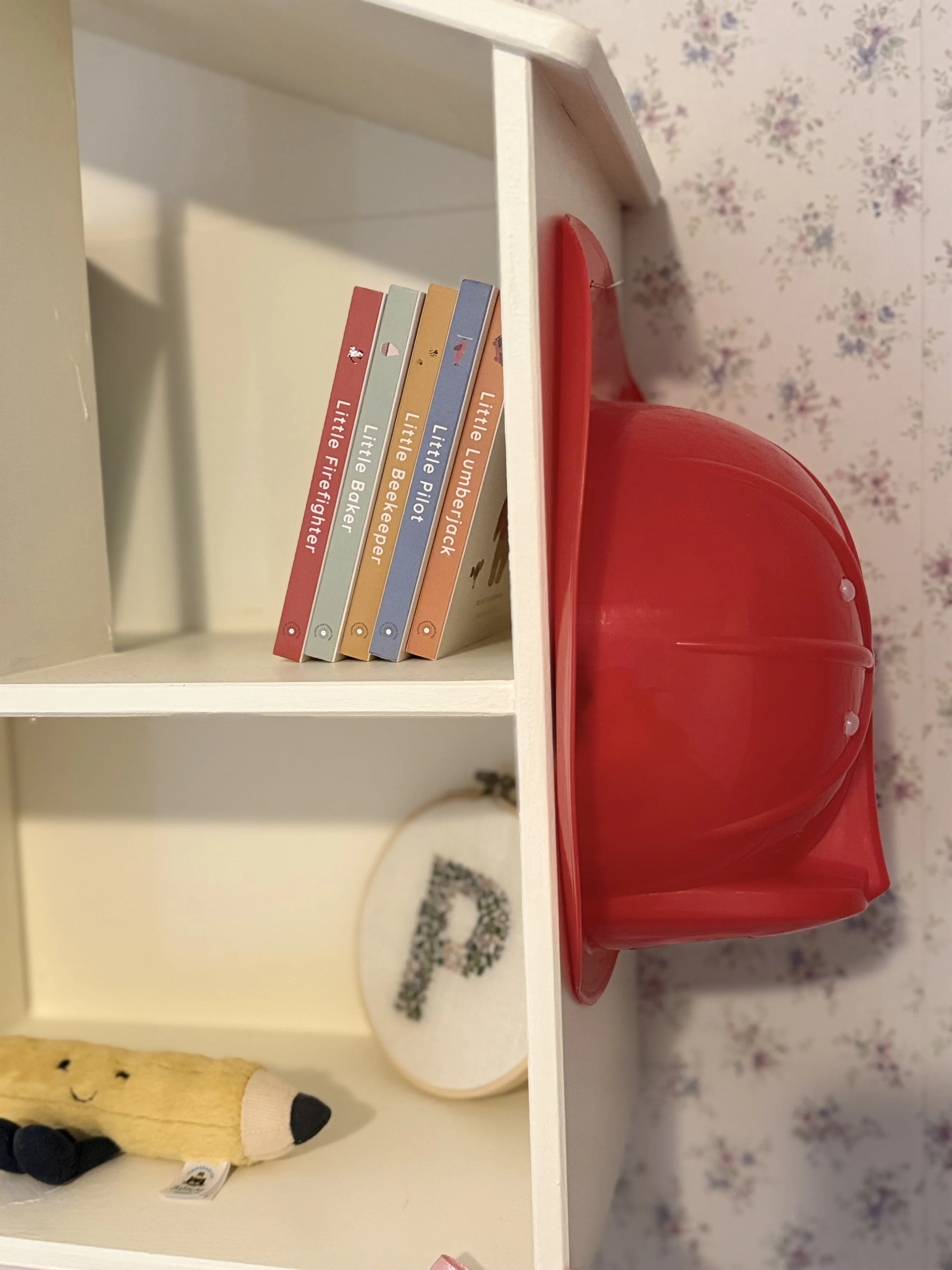 A white shelf with a row of small colorful books, a red container, a plush toy, a firefighter helmet and some decorative items against a floral wallpaper background.