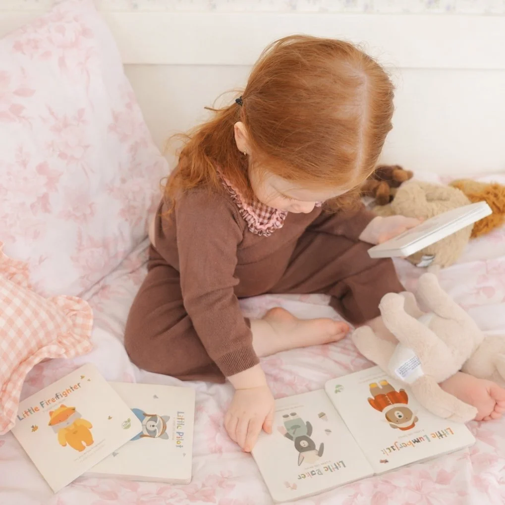A young girl with red hair, wearing a brown outfit, reading picture books and playing with stuffed animals on a pink floral bedspread.
