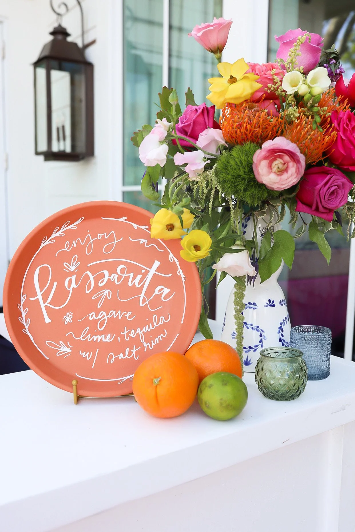 A table with a floral arrangement in a white vase, a pink decorative plate with white text, two oranges, one green apple, and small candles, set outdoors near a house.