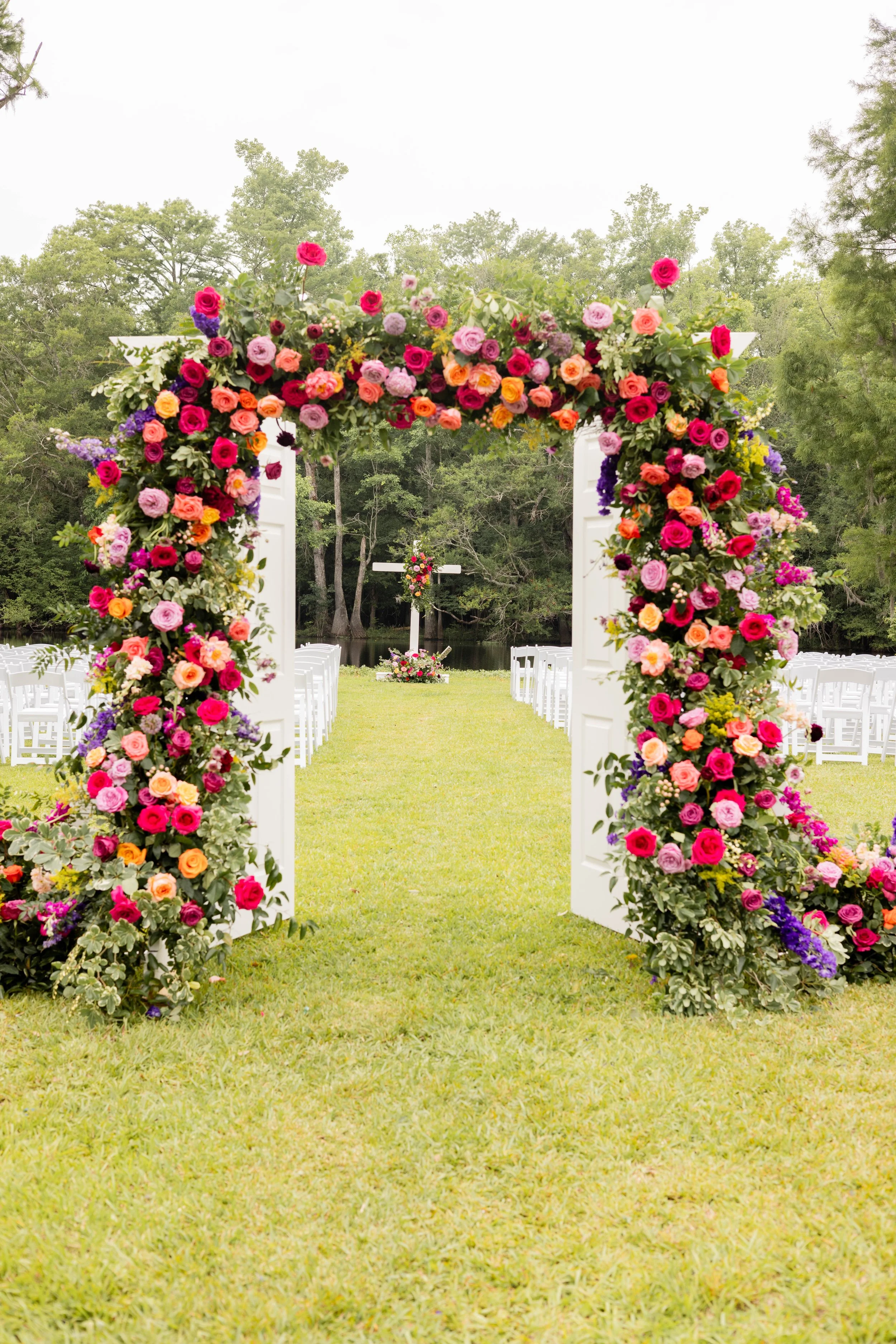 Decorated outdoor wedding arch with colorful flowers, leading to a cross at a ceremonial altar in a grassy area surrounded by trees.