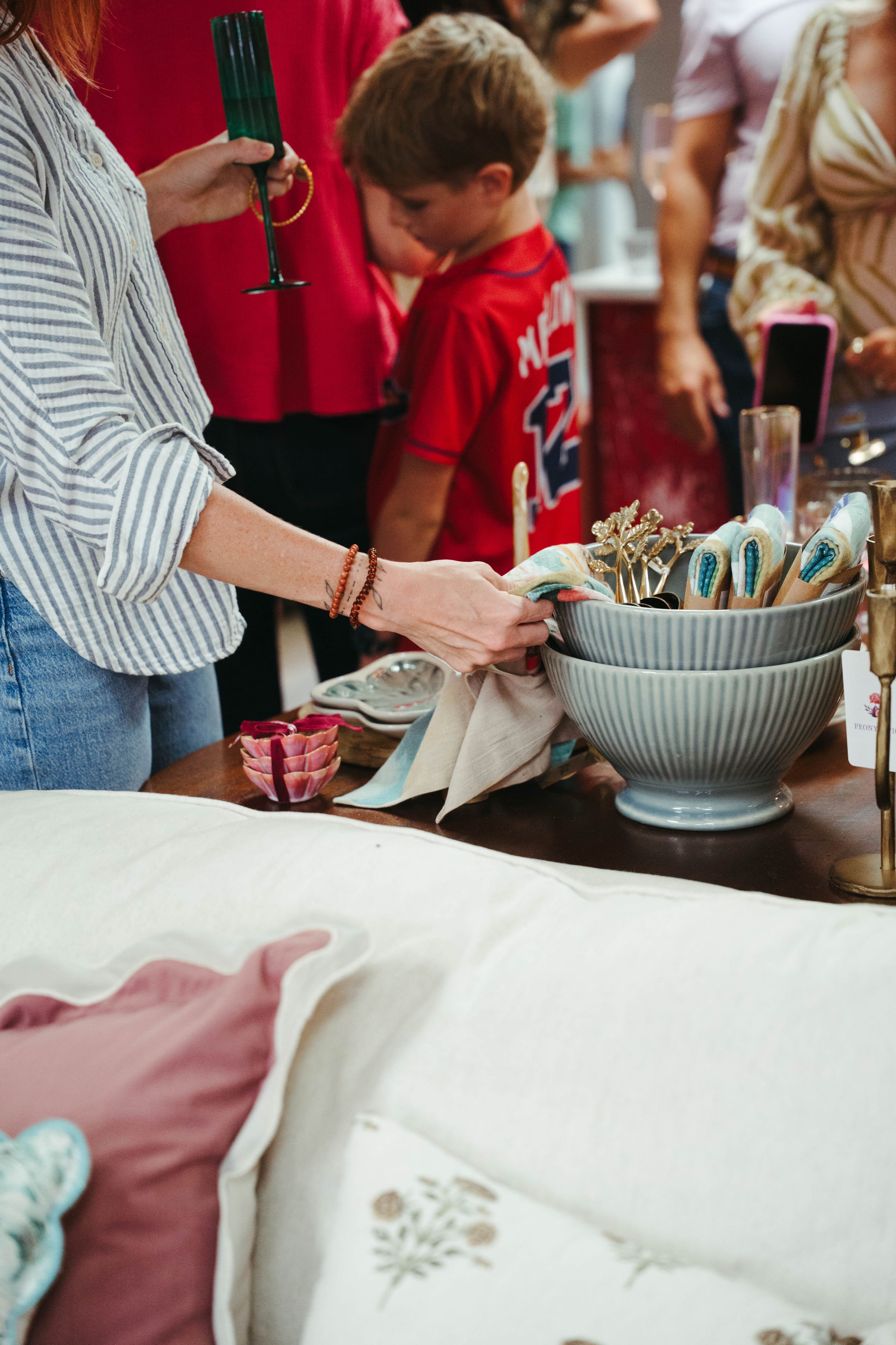 People shopping or browsing at a marketplace or craft fair, with a focus on a woman reaching into a bowl of decorative utensils on a table.