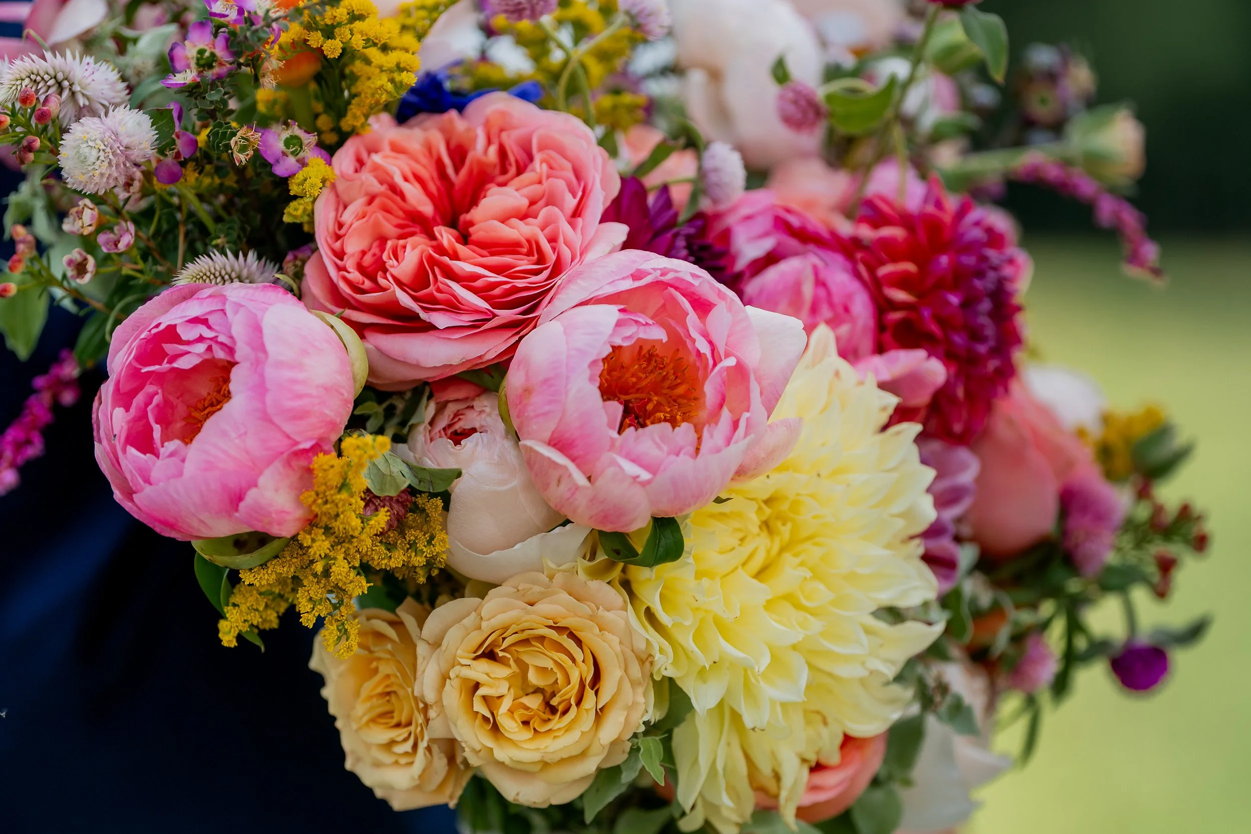 Colorful bouquet of various flowers including pink peonies, yellow dahlias, and other vibrant blossoms, with a blurred green background.