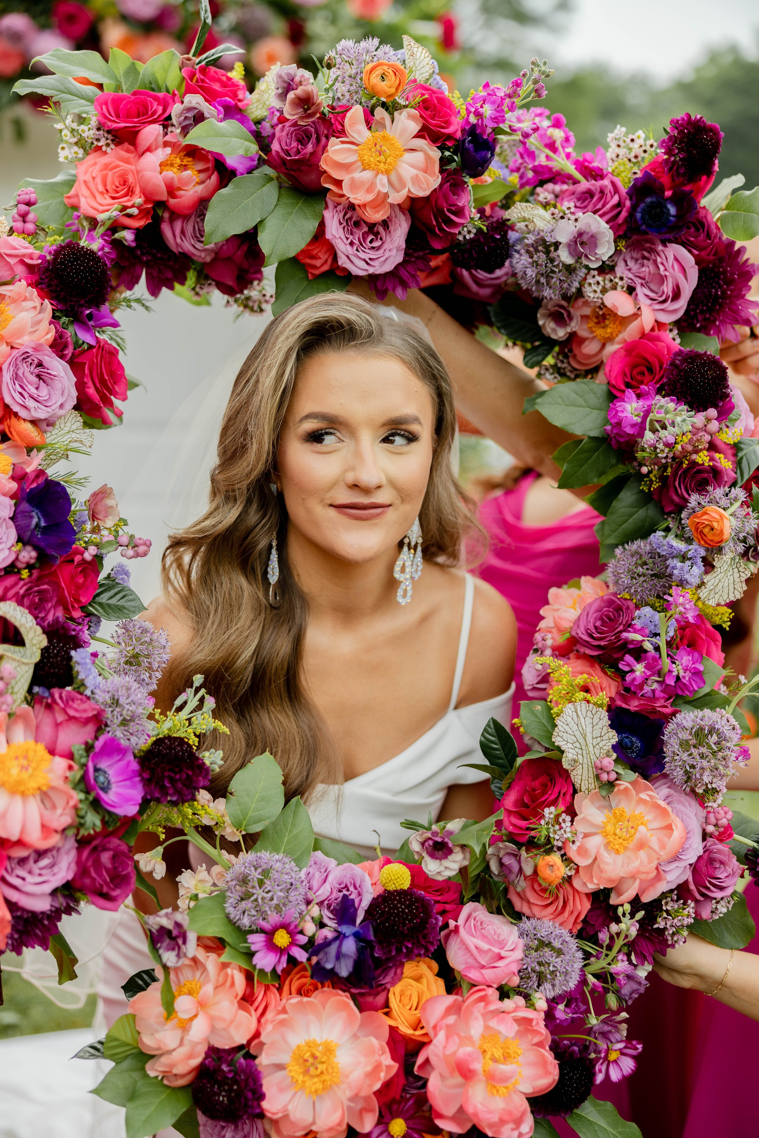 A woman with long wavy brown hair and earrings, surrounded by colorful flowers, holding a floral wreath, at a wedding or celebration event.
