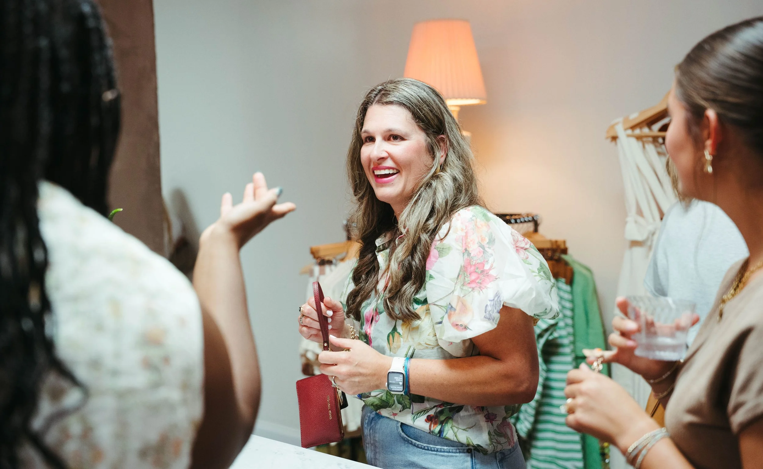 A group of women socializing indoors, with one woman smiling and engaging in conversation, wearing a floral blouse and holding a wallet.