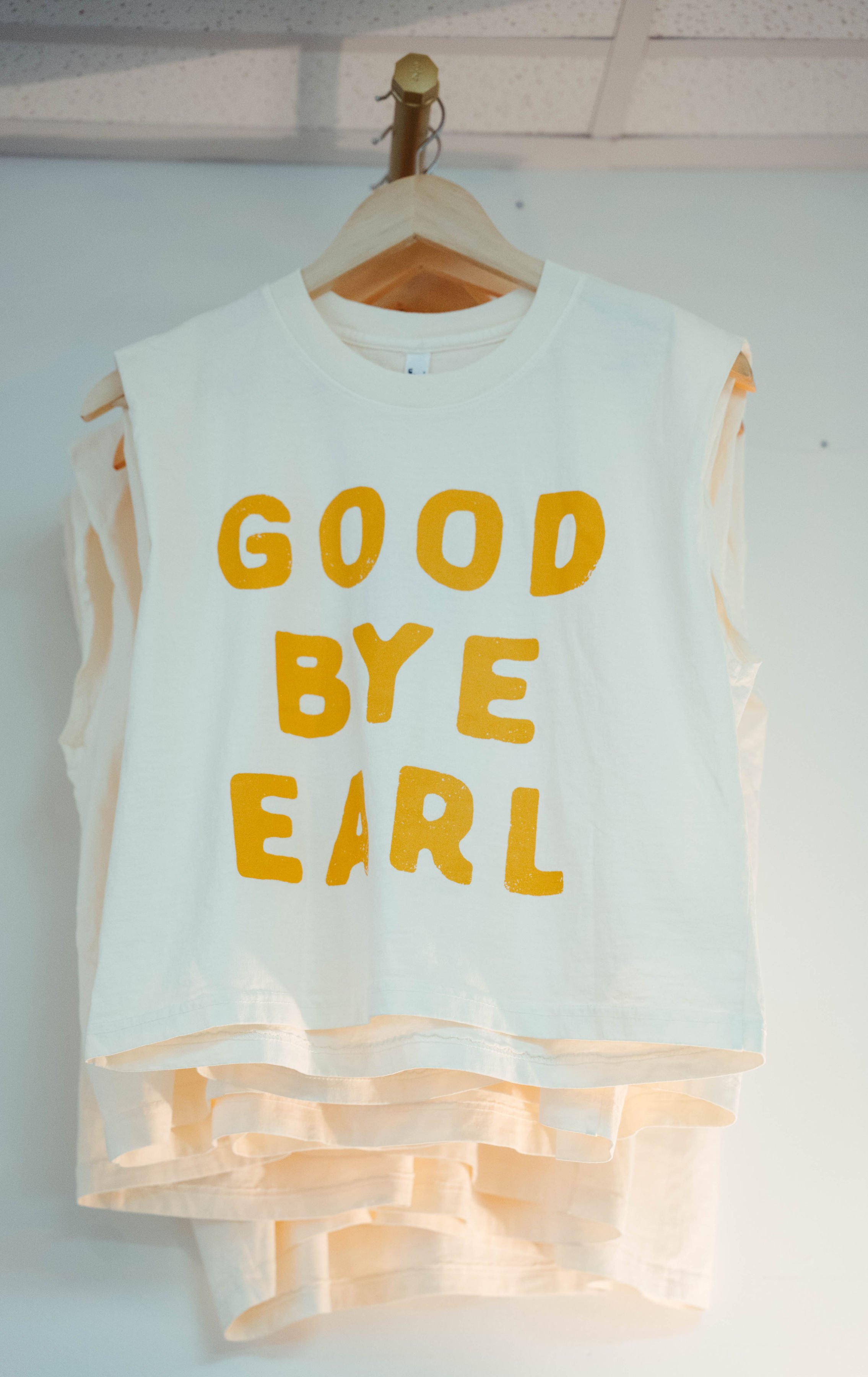 A sleeveless white shirt with the words "GOOD BYE EARL" printed in yellow text, hanging on a wooden hanger.