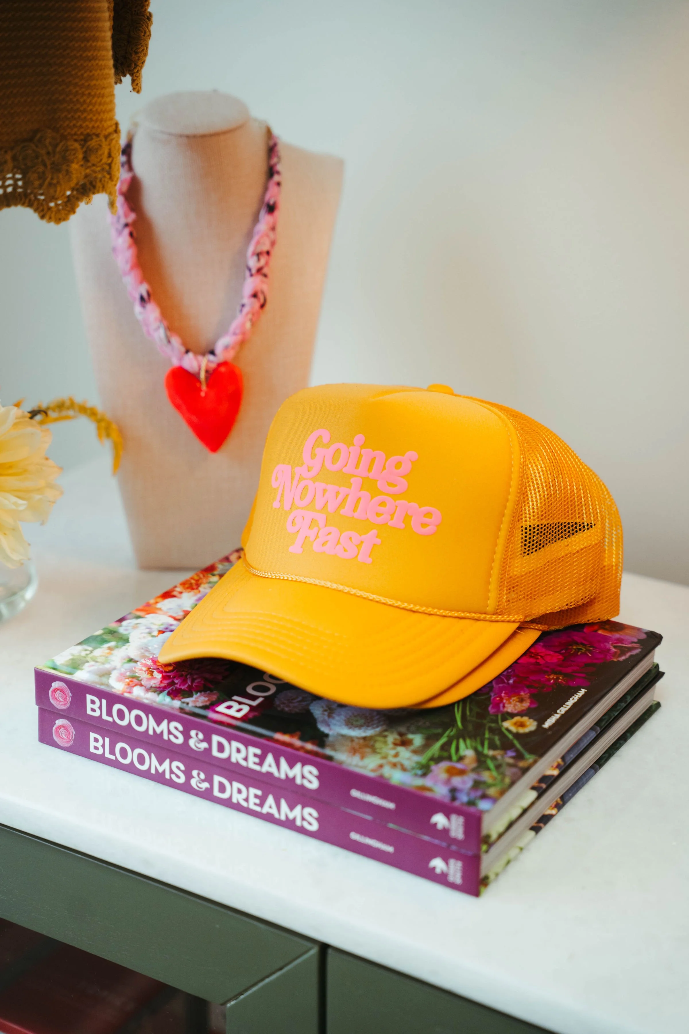 A yellow trucker-style cap with pink text reading 'Going Nowhere Fast' on top of two hardcover books titled 'Blooms & Dreams'. The books have colorful flower covers. In the background, there is a necklace with a large red heart pendant displayed on a