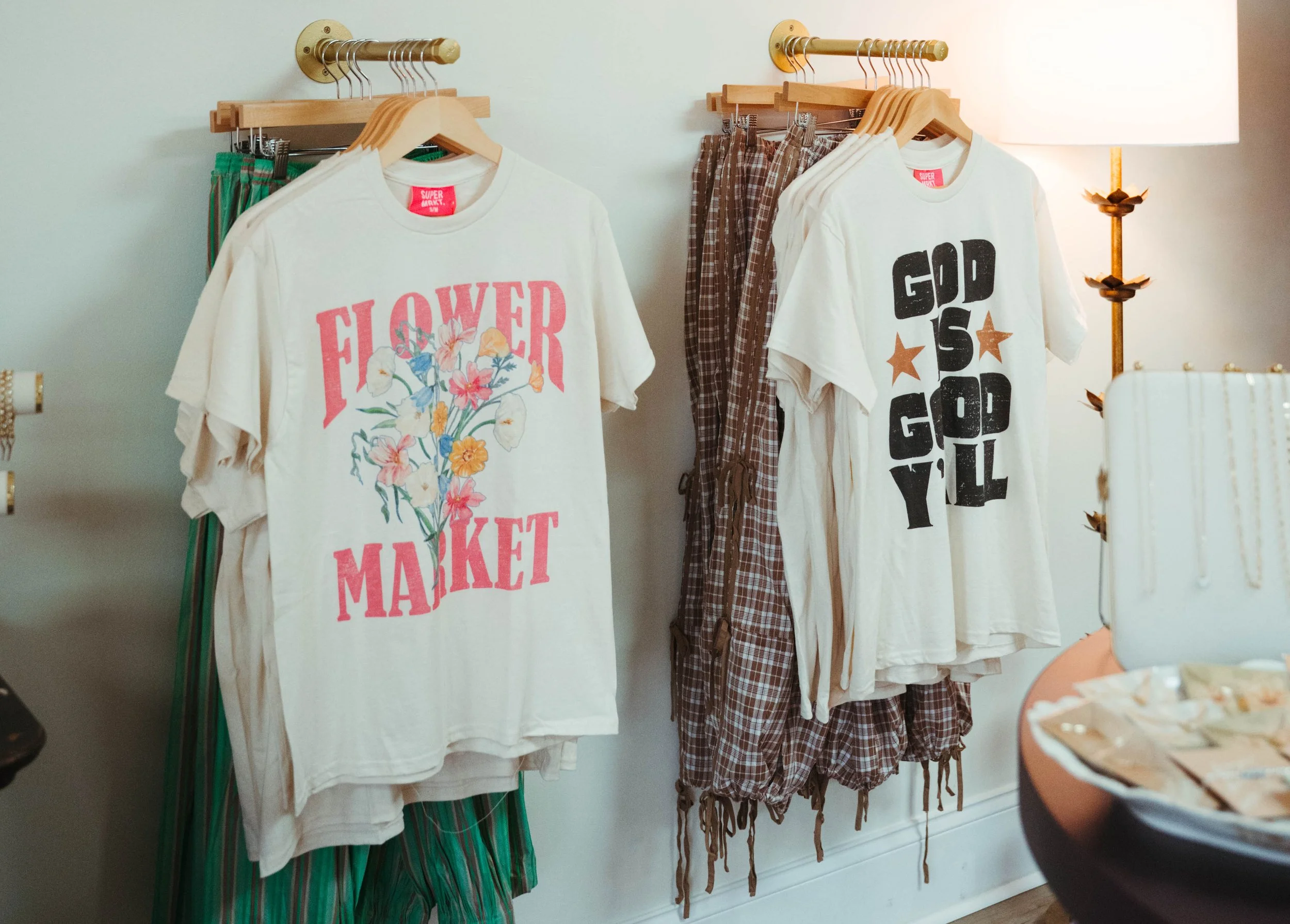 Display of casual clothing with graphic prints hanging on a wall. Two white T-shirts, one with "FLOWER MARKET" and a floral design, and the other with "GOD IS GOD Y'ALL" and star graphics. Behind the shirts, plaid shorts and pants are visible.