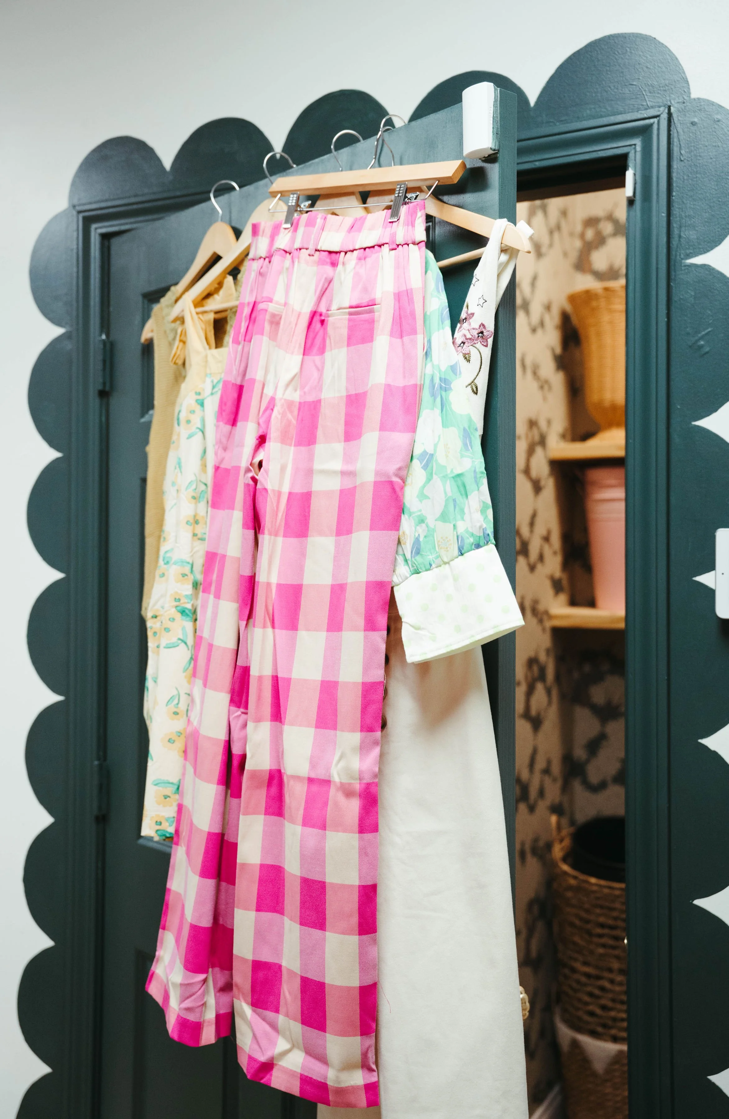 Clothes hanging on a closet door with decorative scalloped trim, inside a room with floral wallpaper and shelves with baskets.