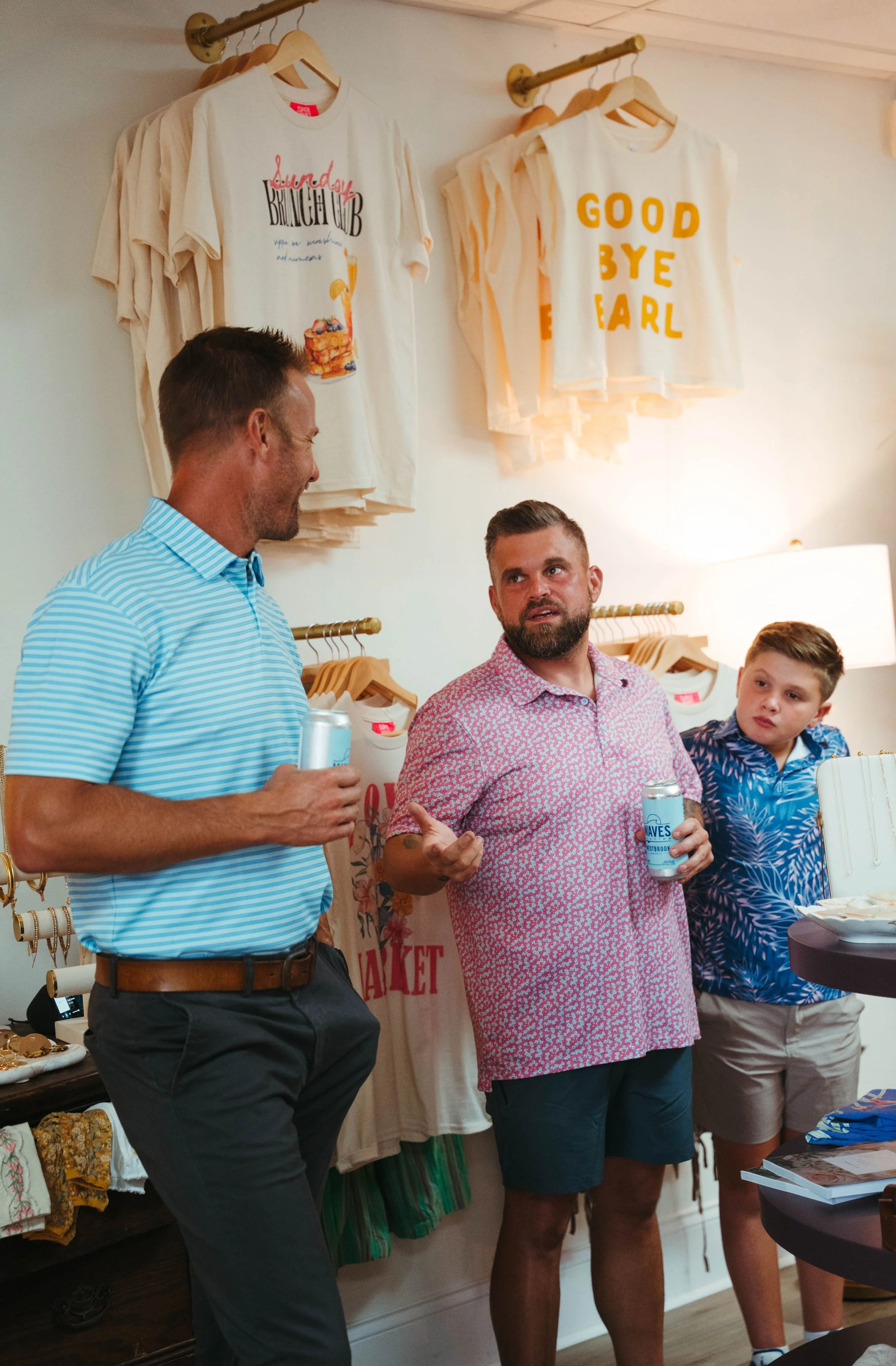 Three men standing and talking in a store with t-shirts hanging on the wall; two of them hold cans of drinks.