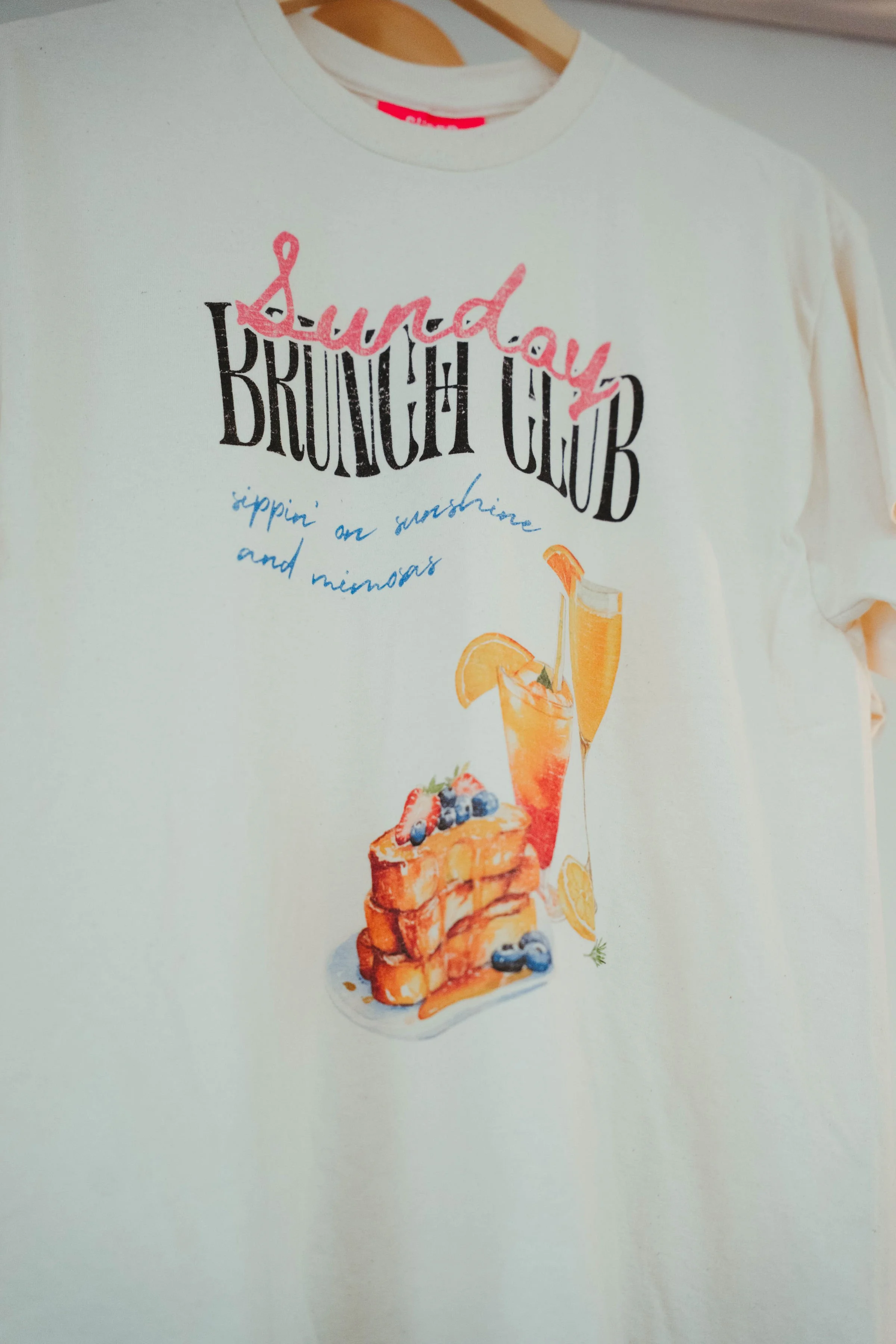 White T-shirt with graphic design featuring stacked waffles, blueberries, and a mimosa, with playful text above that reads 'Sound, Brunch Club' and 'sippin' on sunshine and mimosas'.