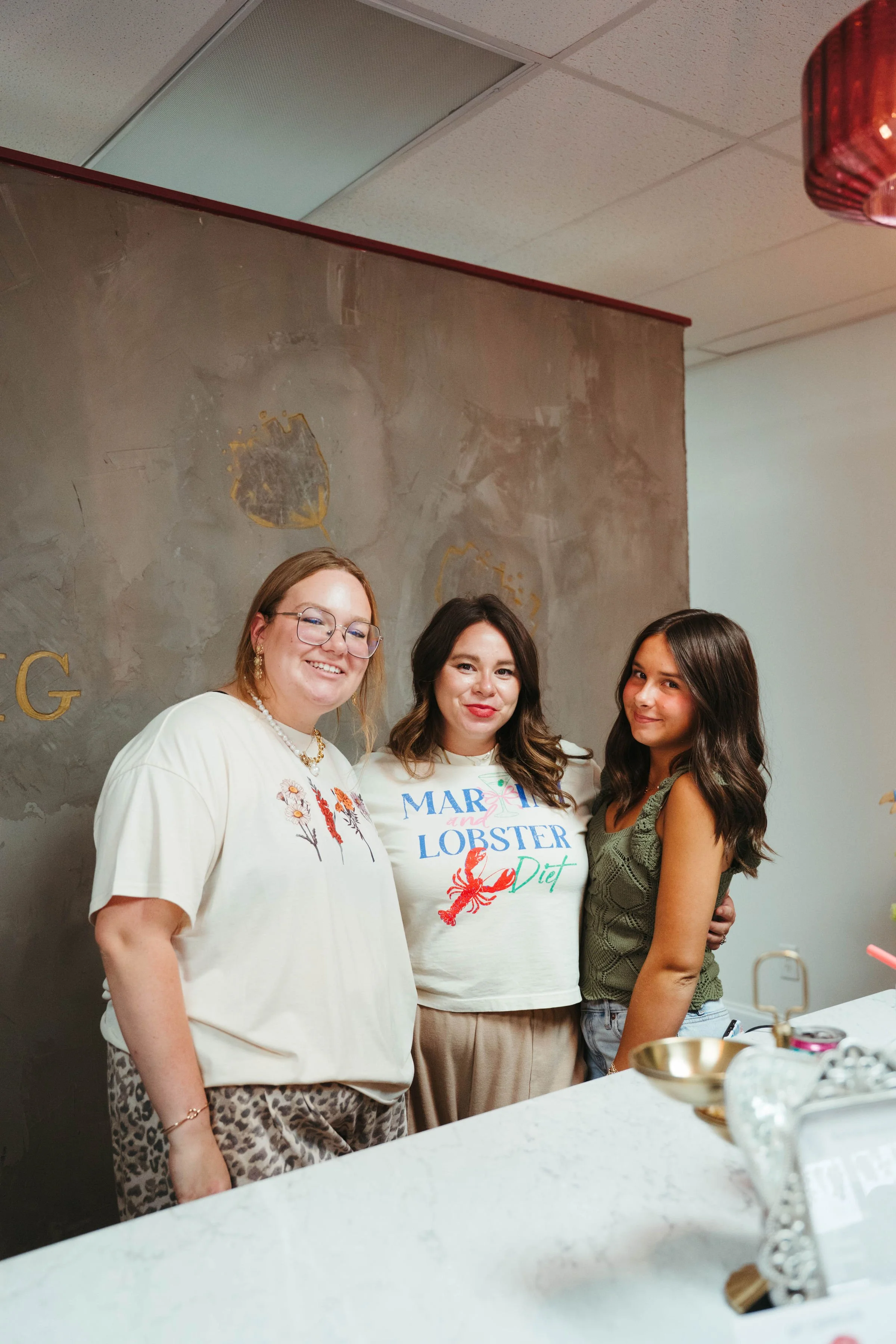 Three women standing together indoors, smiling at the camera, with a textured gray wall behind them.