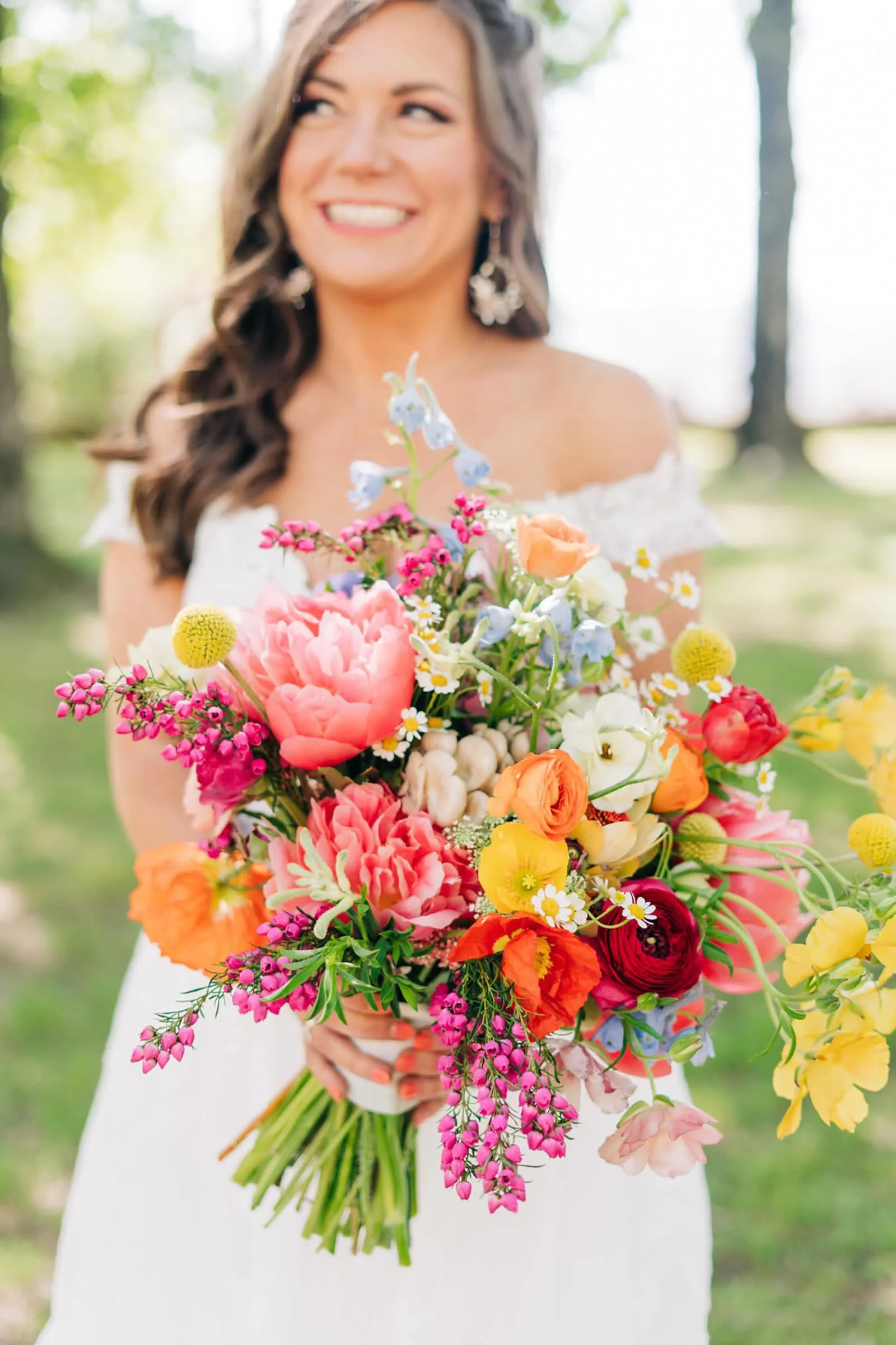 A woman in a white off-shoulder dress holding a colorful bouquet of various flowers outdoors.