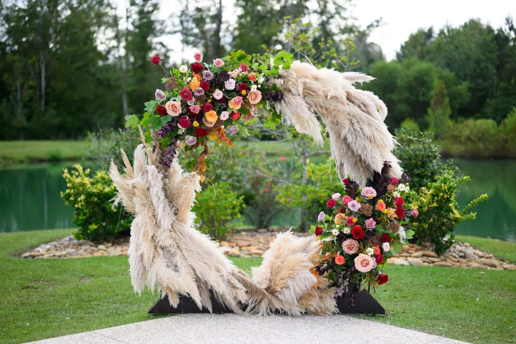 A floral arch made of pampas grass and colorful roses, set outdoors near a body of water and surrounded by greenery.