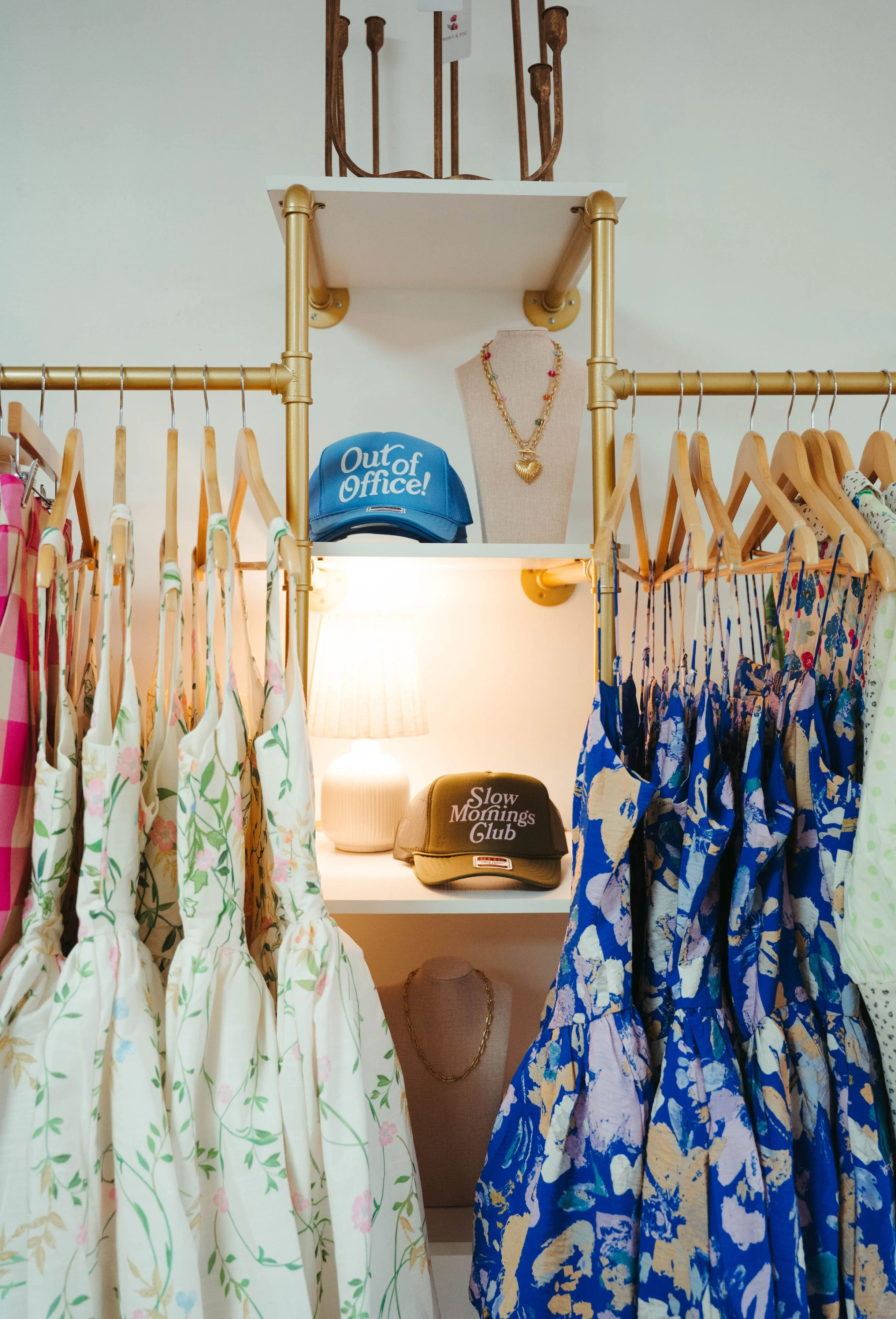 Clothing store display with floral dresses on hangers, a blue 'Out of Office' cap, a 'Slow Mornings Club' cap, a mannequin necklace, and jewelry on a mannequin bust, with a lamp in the background.