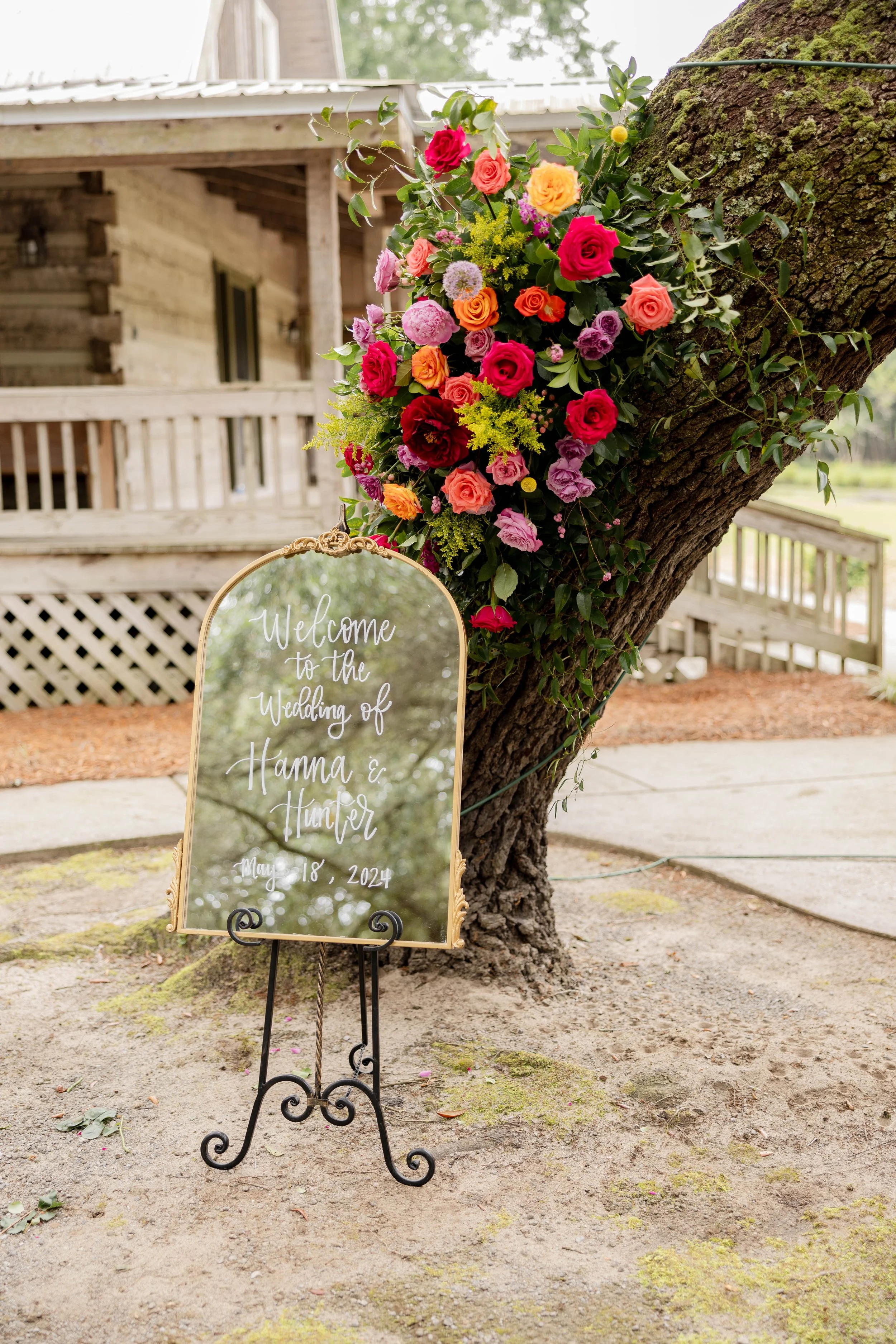 Wedding welcome sign with the text 'Welcome to the wedding of Hanna & Hunter, May 18, 2024' displayed on a mirror, standing on a decorative black iron easel, next to a large tree with colorful bouquet of pink, red, orange, purple, and yellow flowers.