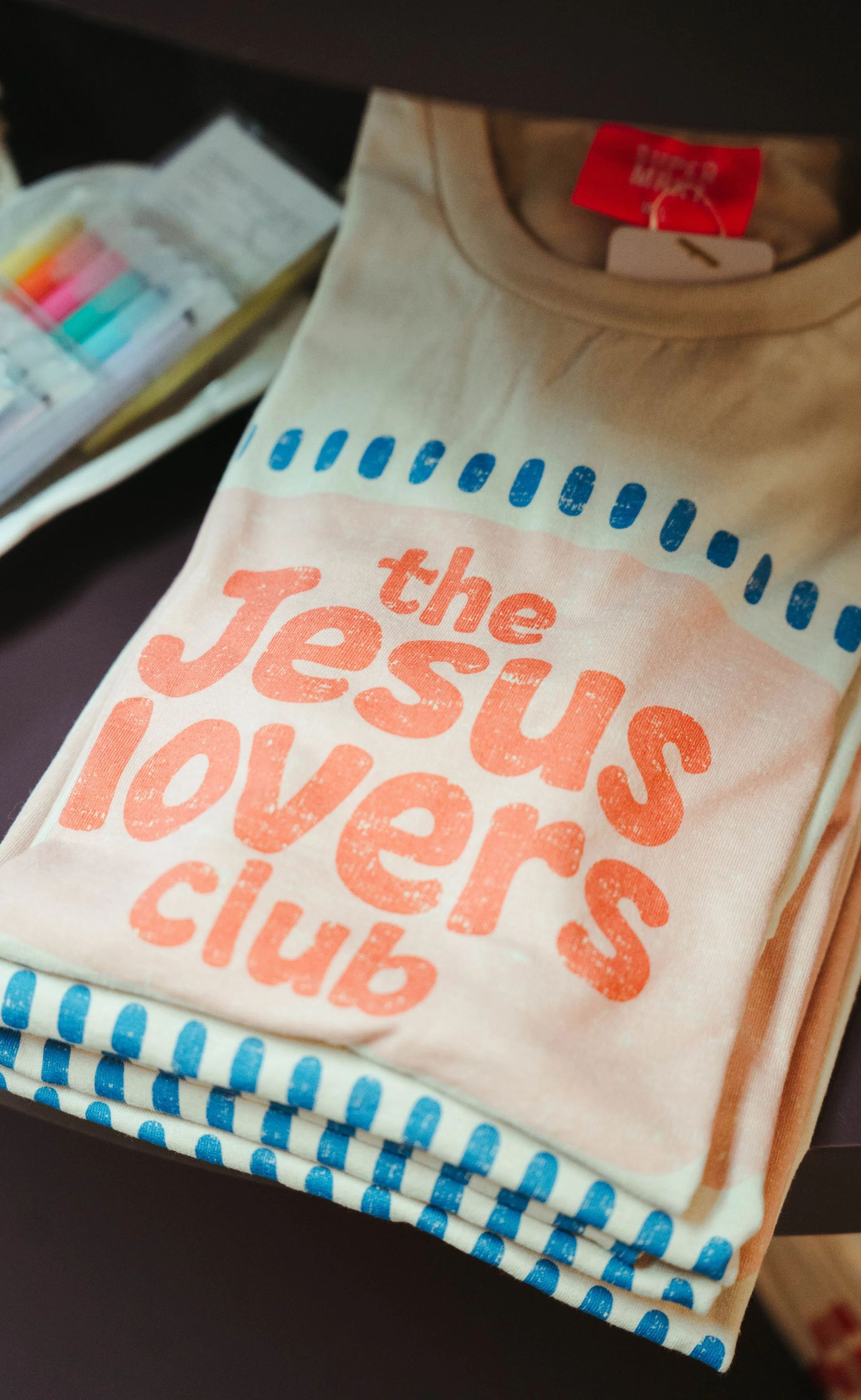 Folded T-shirt with the text 'the Jesus lovers club' printed on it in pink and orange letters. The shirt is beige and is partially visible with blue and white patterned fabric underneath. There is a pack of colored highlighters or markers in the back