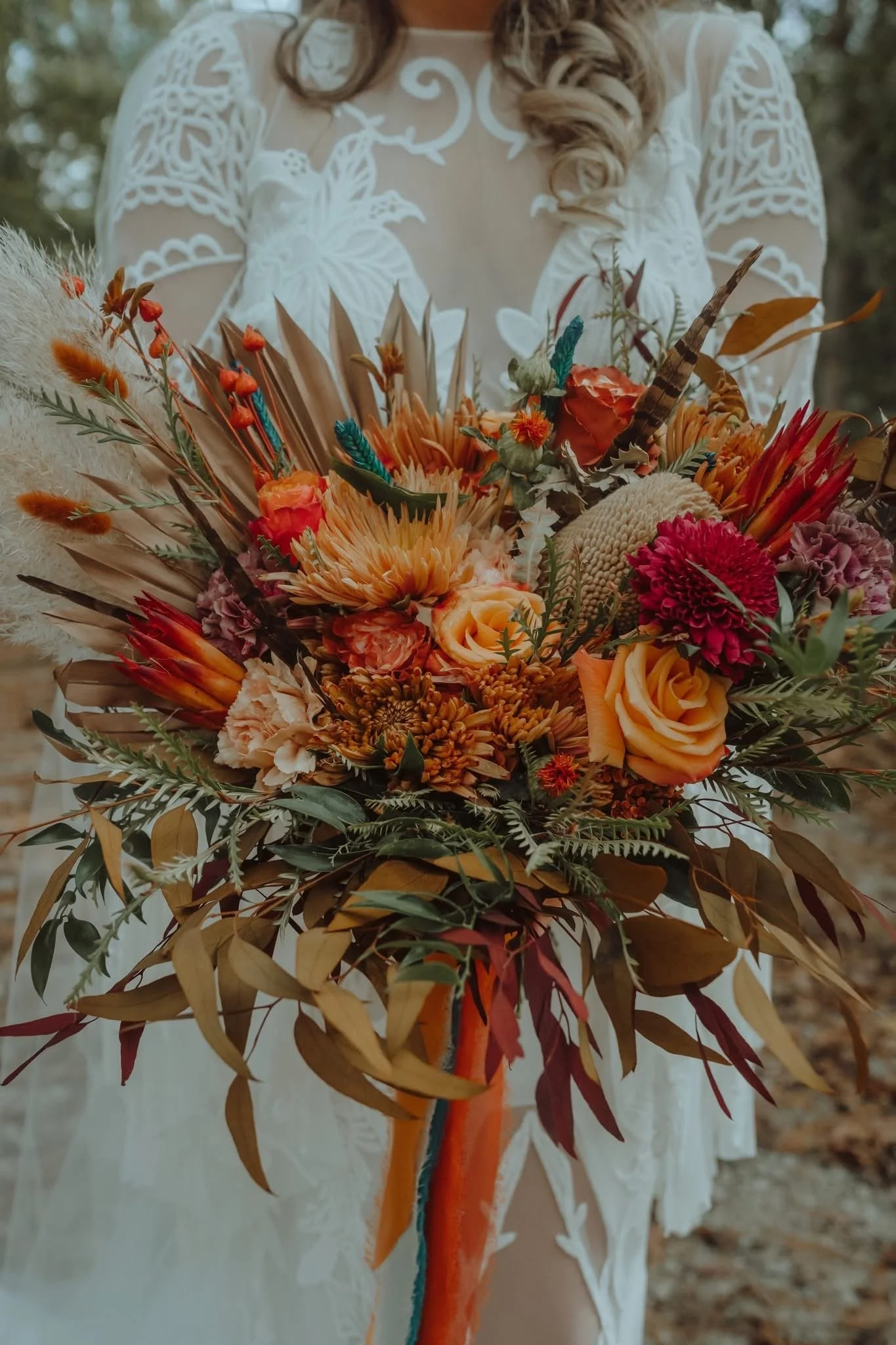 Close-up of a woman in a white lace dress holding a large, colorful bouquet of flowers, including roses, chrysanthemums, and dried foliage, outdoors with blurred background.