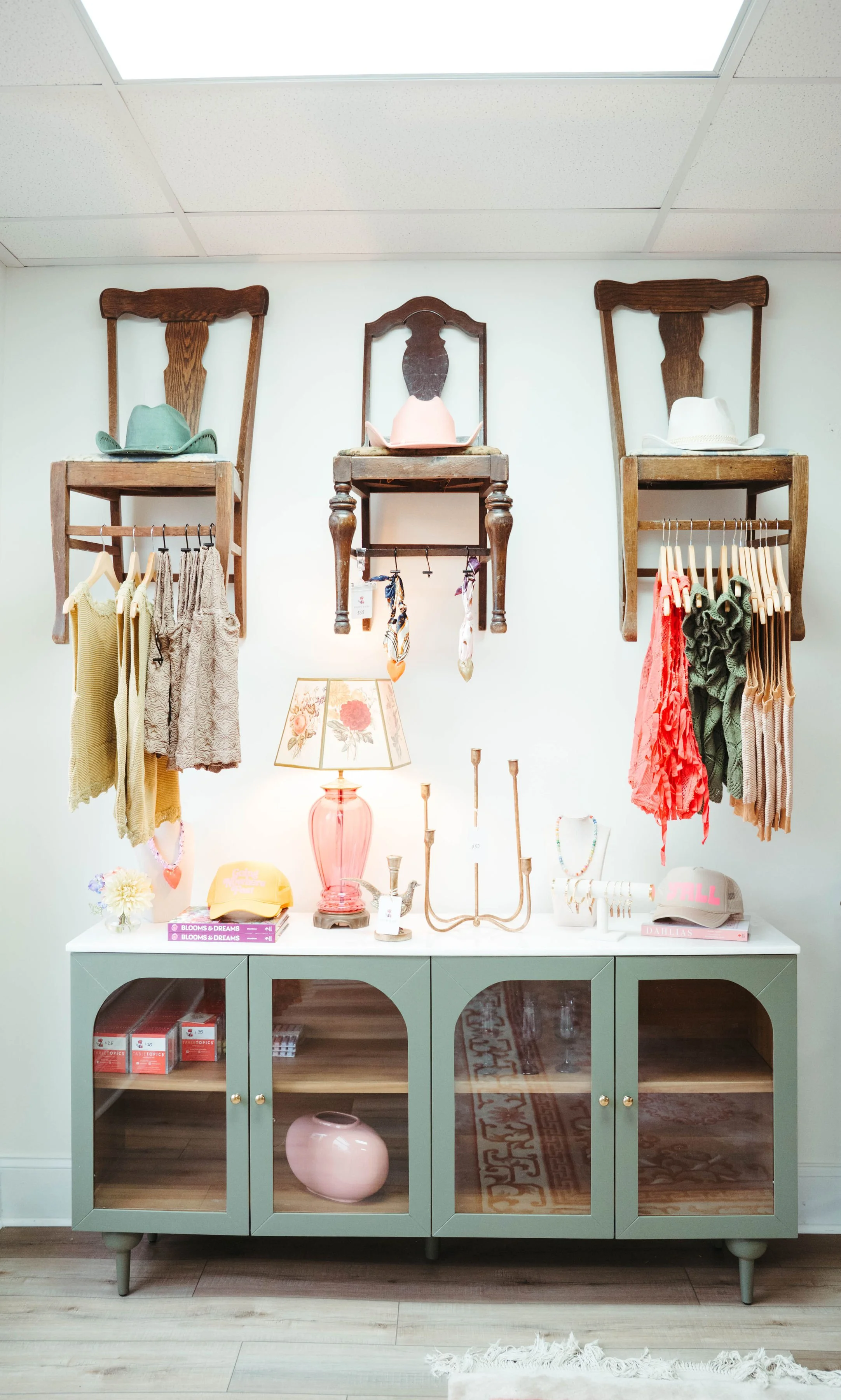 A display with vintage decor, hats, and clothing on wooden chairs mounted on a wall above a pastel green cabinet. The cabinet has glass doors and contains books and decorative objects. A lamp with a floral shade, jewelry, and small decorative items a
