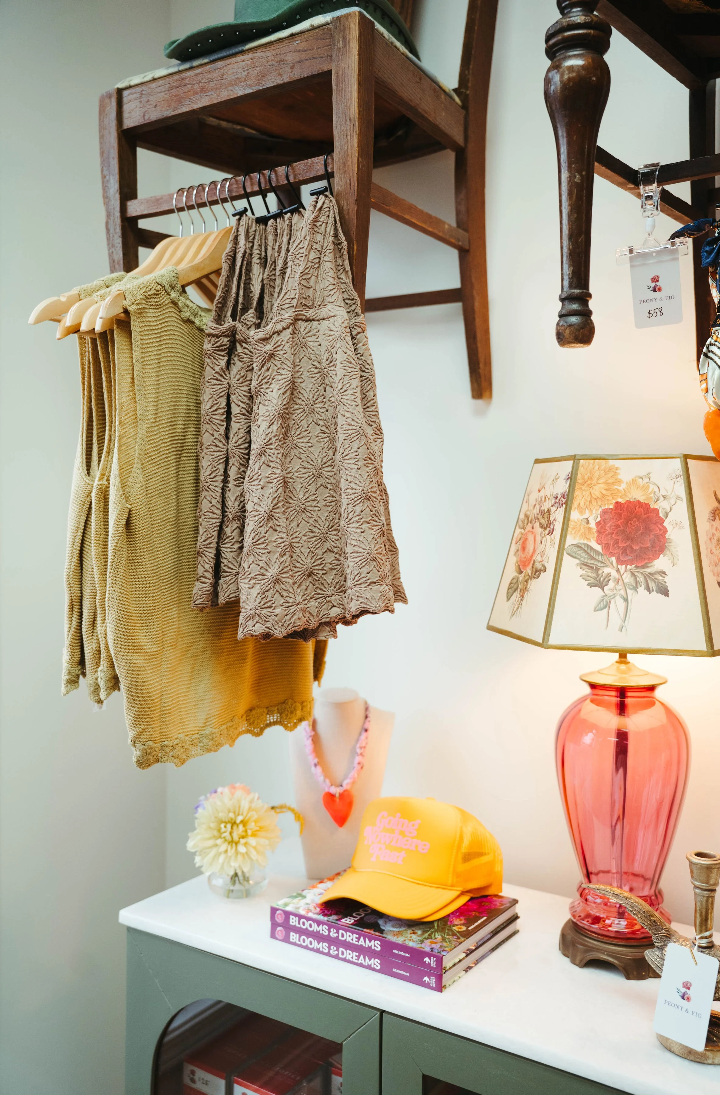 Display of clothing items, including yellow and patterned skirts or shorts, hanging on black hangers from a wooden rack. A pink lamp with a floral shade, a small flower arrangement, a yellow cap, books, and a necklace mannequin are also on a white ca