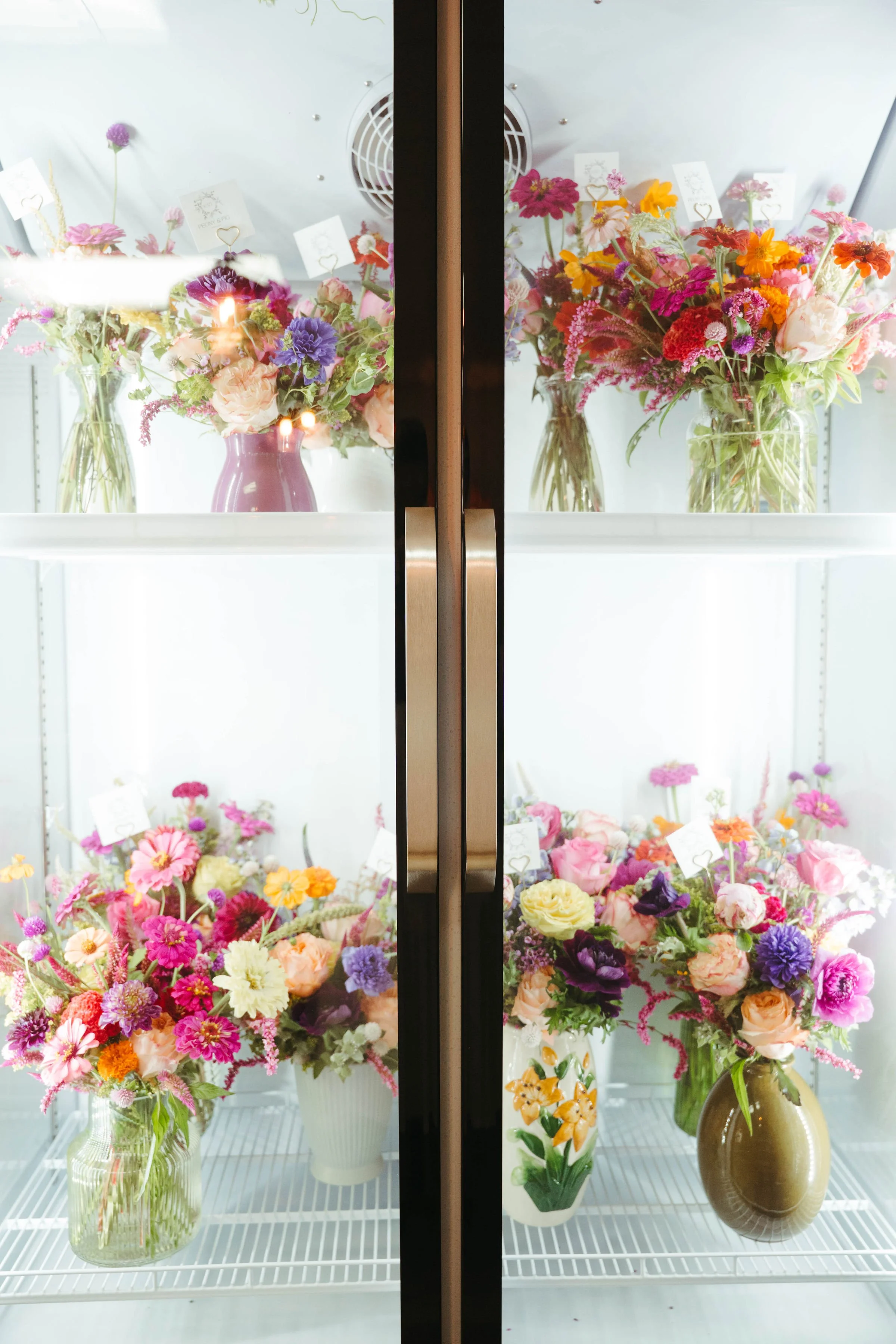 Display of colorful flower arrangements in glass and ceramic vases inside a refrigerated showcase.