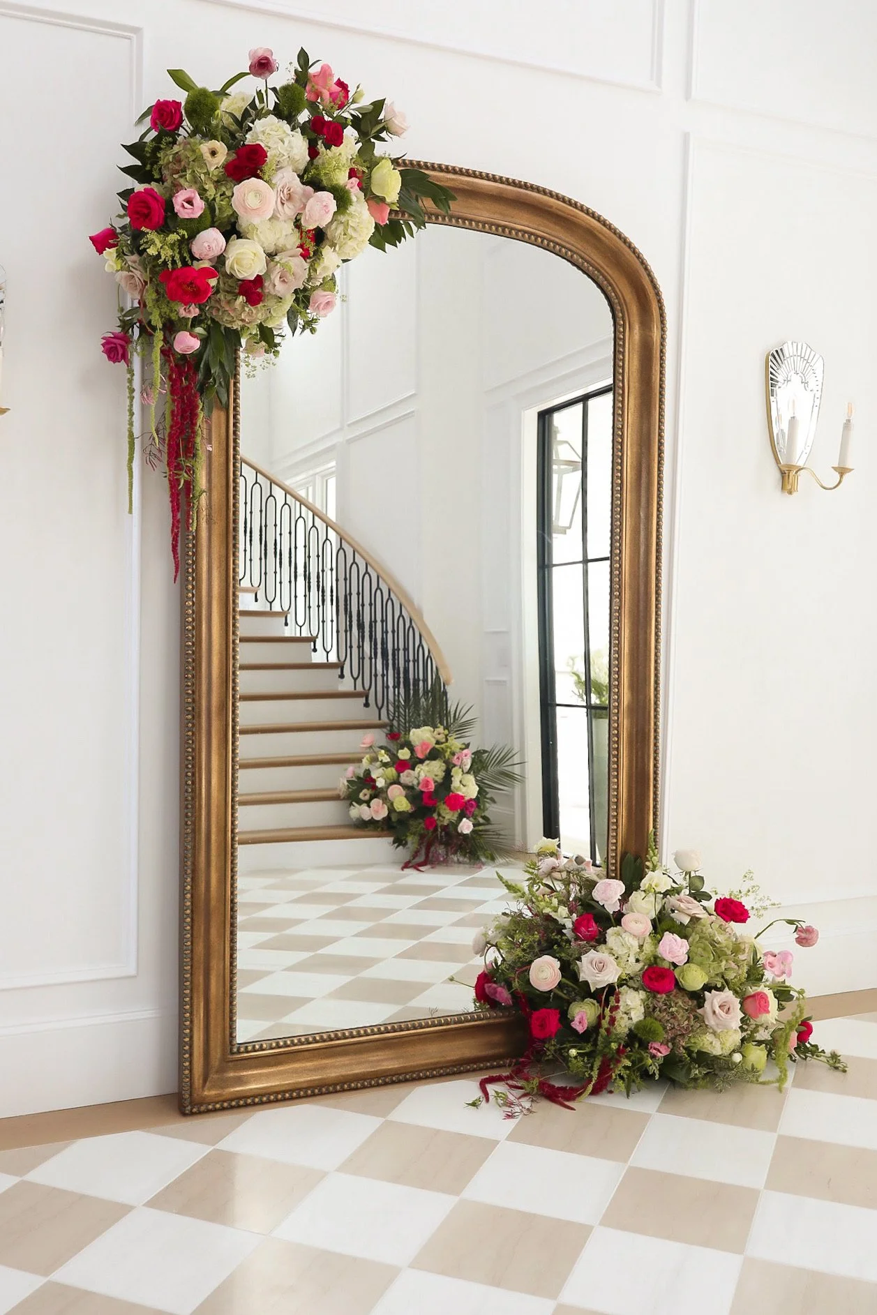 Decorated gold-framed mirror in a bright, elegant room with floral arrangements in pink, white, and red, reflecting staircase and window.