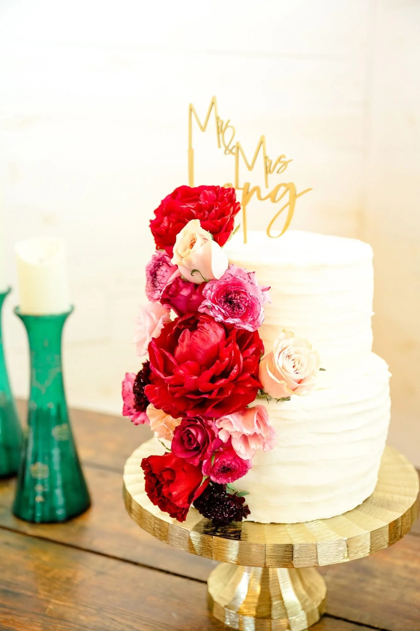 A wedding cake decorated with an arrangement of pink and red flowers, topped with a "Mr. & Mrs. Loving" gold cake topper, on a gold cake stand.