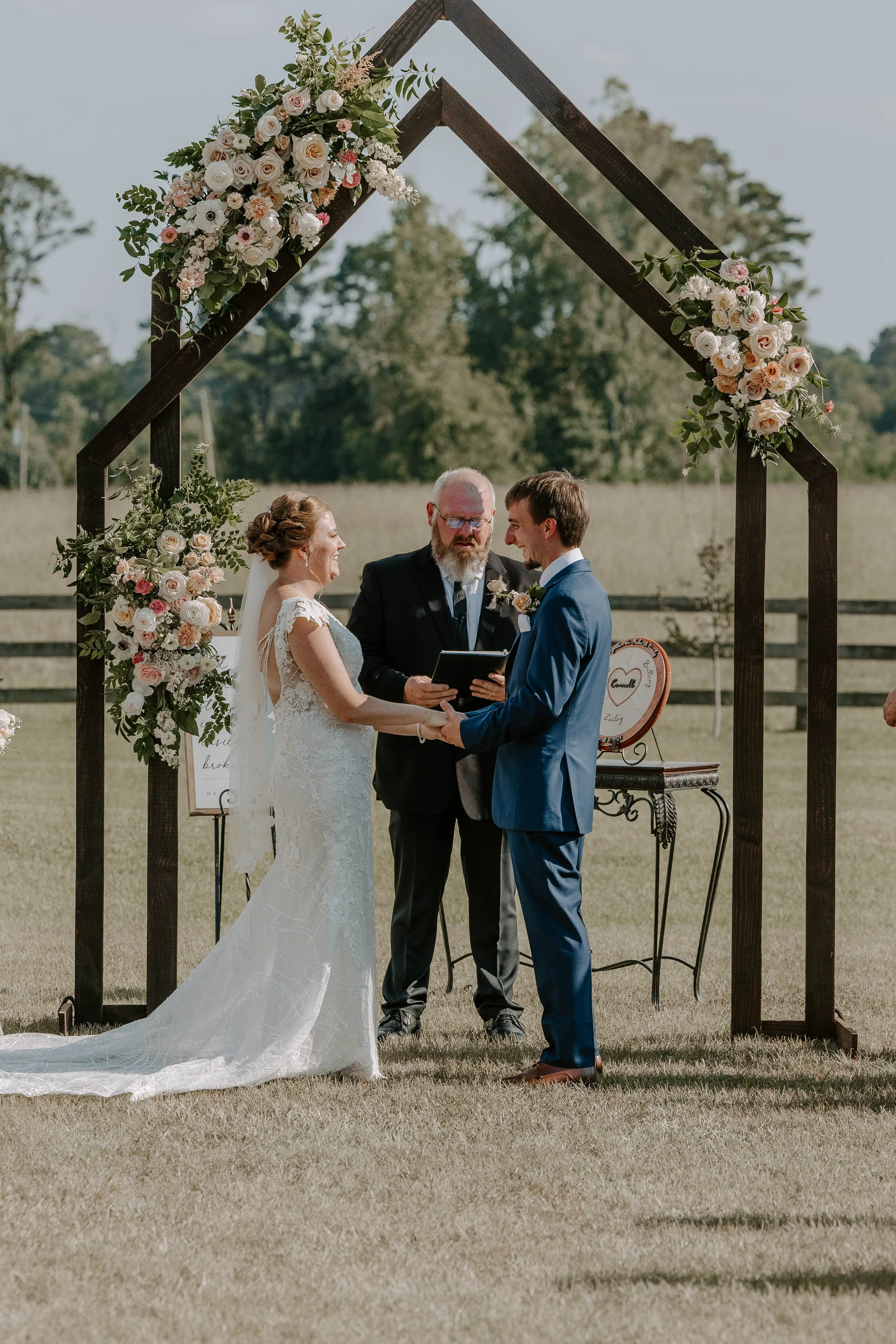 A couple getting married outdoors under a wooden arch decorated with flowers, with an officiant standing between them, holding hands and exchanging vows.
