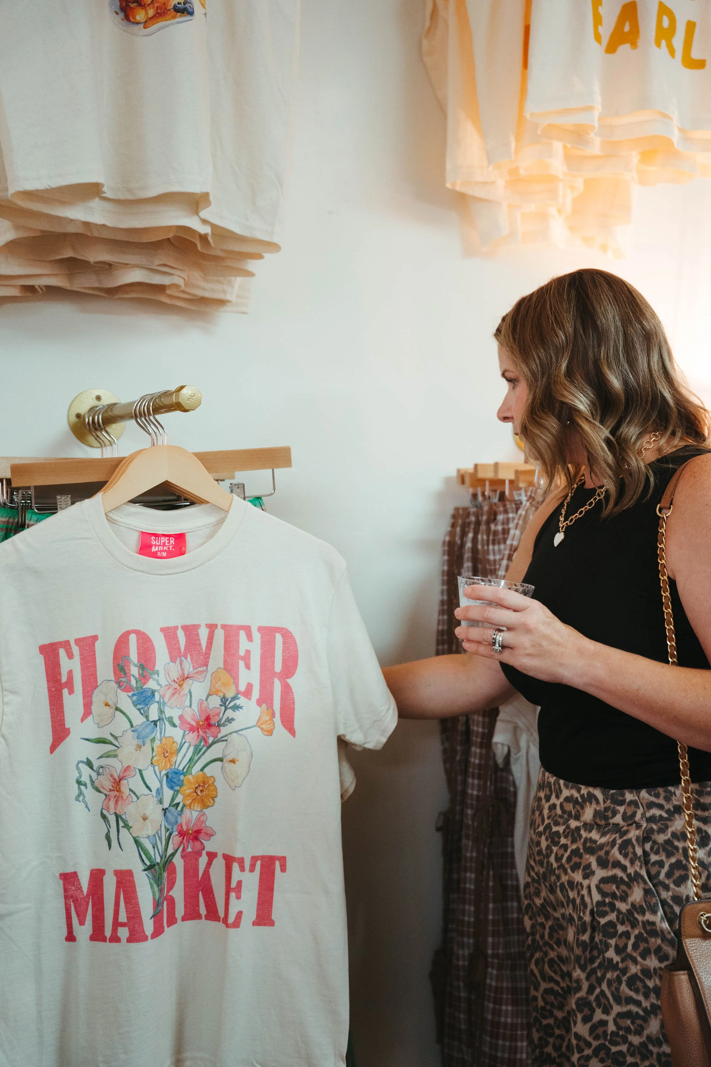 Woman shopping for clothes in a boutique, holding a plastic cup, with a cream t-shirt featuring a floral design and the words "Flower Market" hanging on a rack.