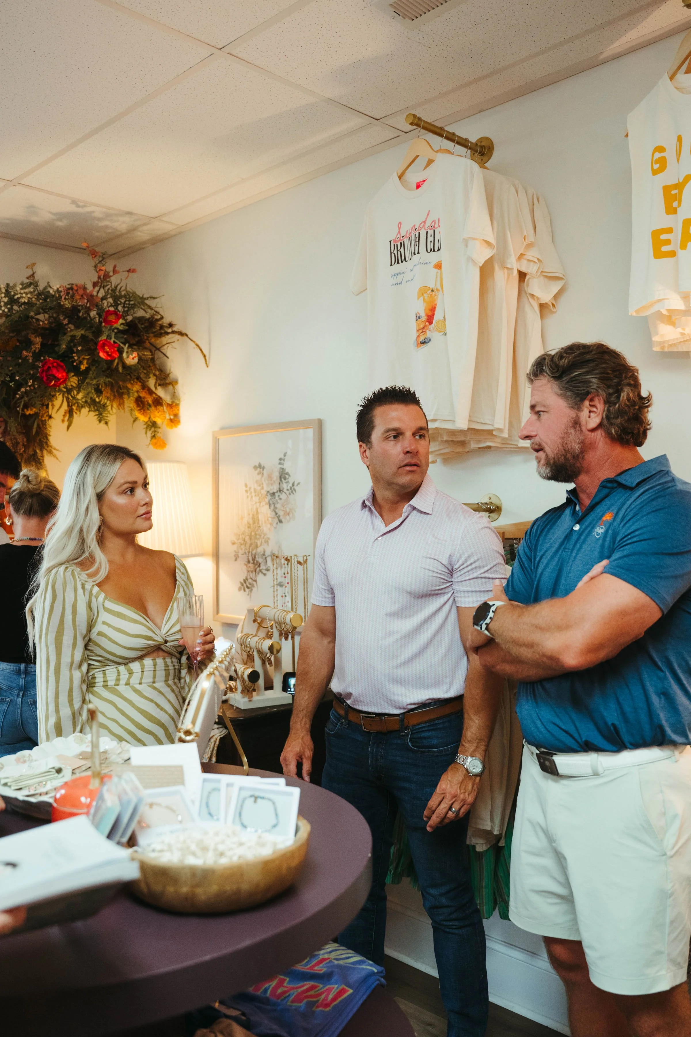Three people having a conversation inside a store, with T-shirts hanging on the wall behind them and a table with merchandise in the foreground.