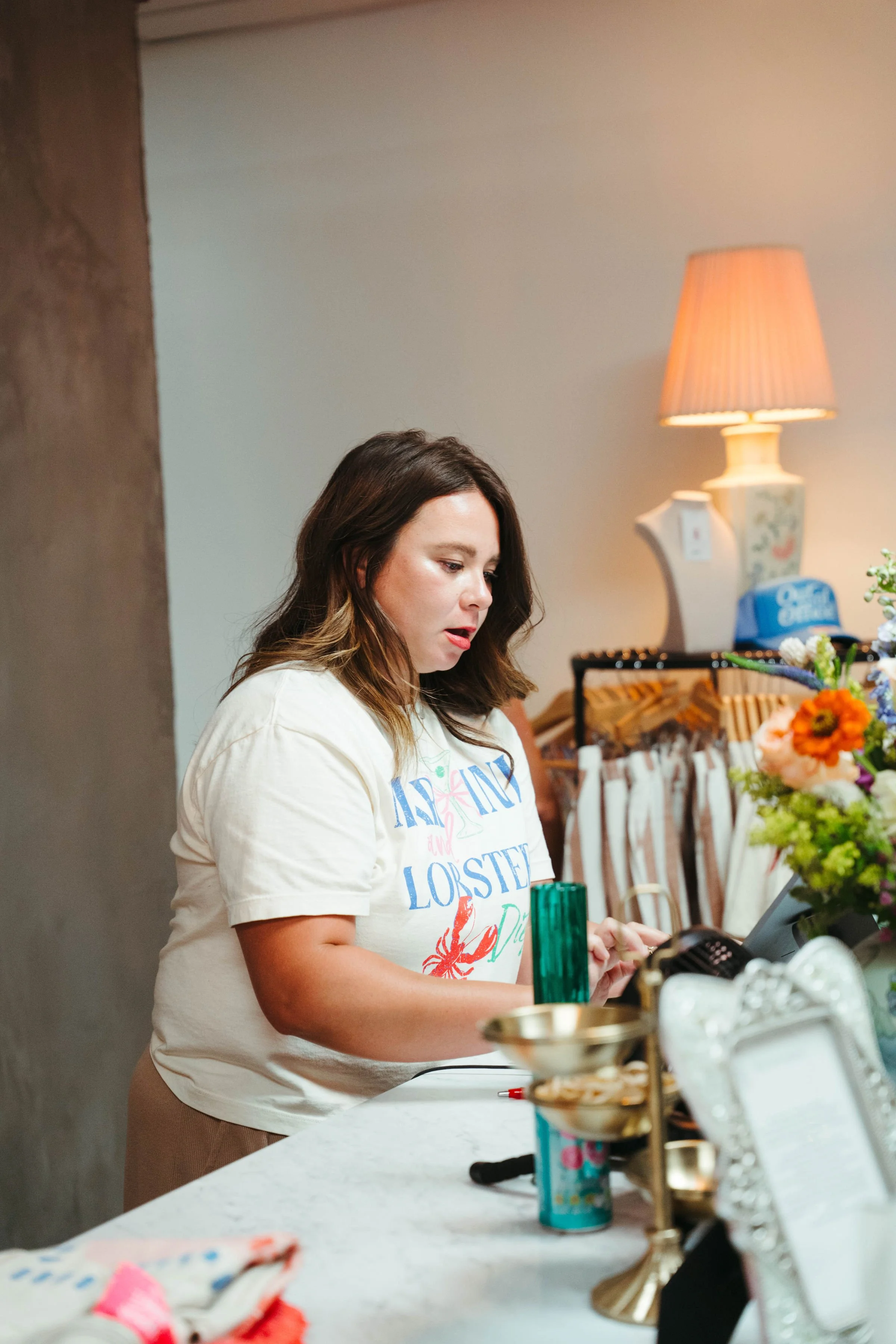 A woman standing behind a white counter, talking on the phone, surrounded by various decorative items and flowers, with a lamp and hanging clothes in the background.
