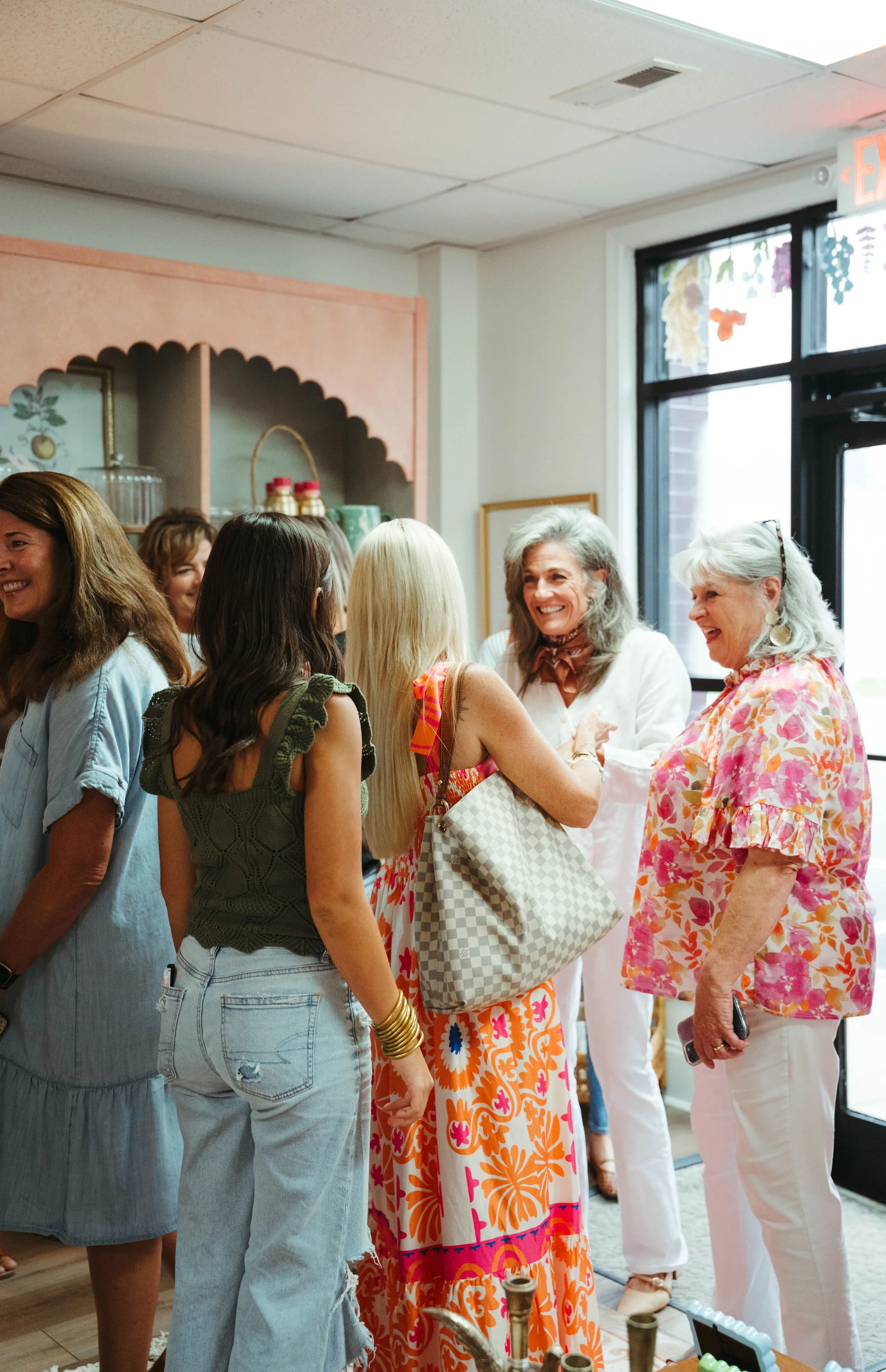 Group of women smiling and chatting inside, with large windows and colorful clothing.