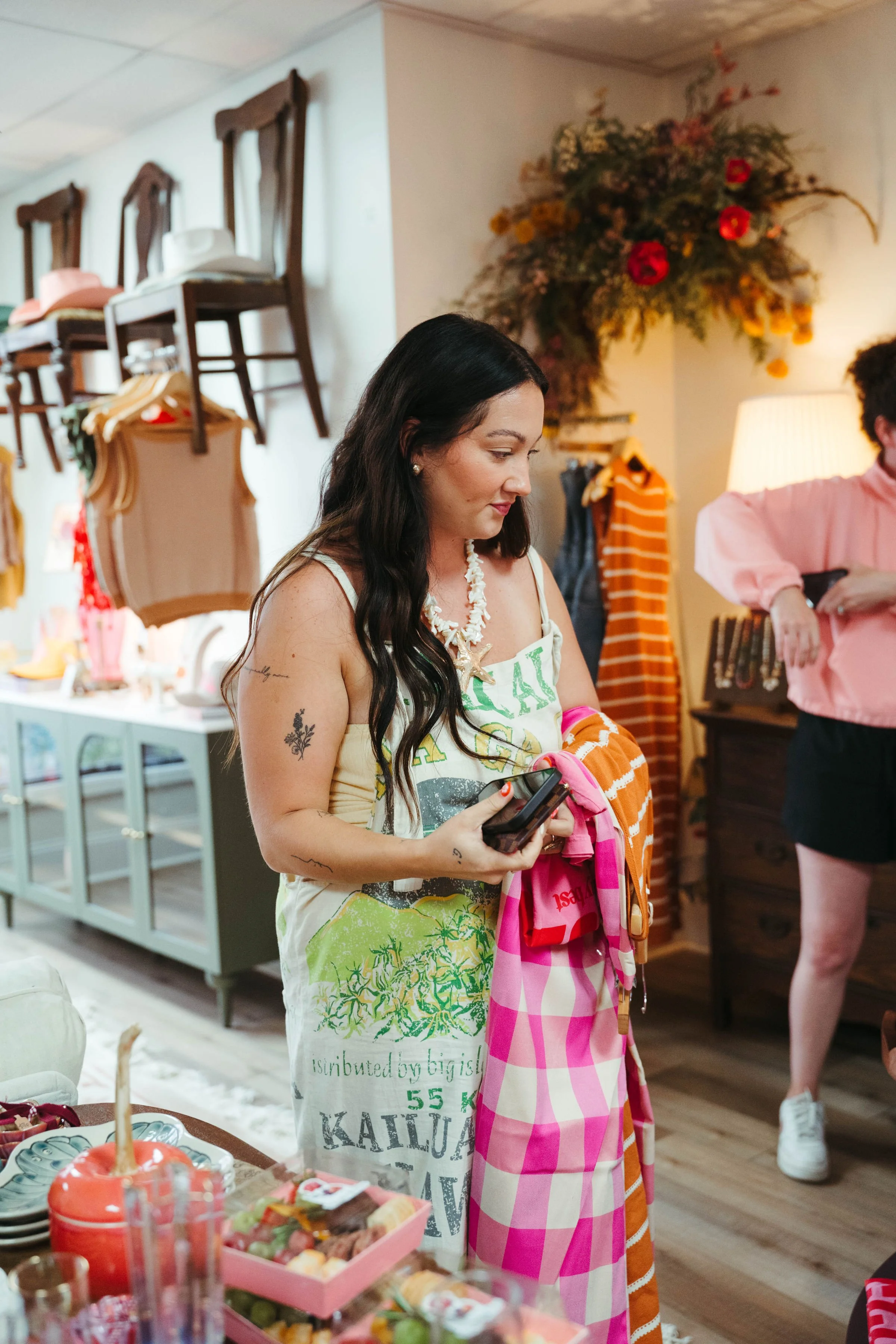 A woman with long dark hair wearing a white dress stands in a boutique store, holding a smartphone and a pink checkered towel. She has tattoos on her arms and is wearing a shell necklace. The store has racks of clothing, decorative items, and a flora