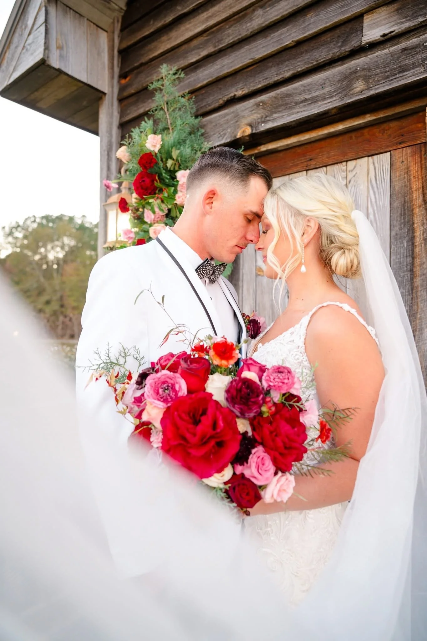 A bride and groom stand with foreheads touching, holding a bouquet of red, pink, and white flowers, in front of a rustic wooden background with floral decorations.