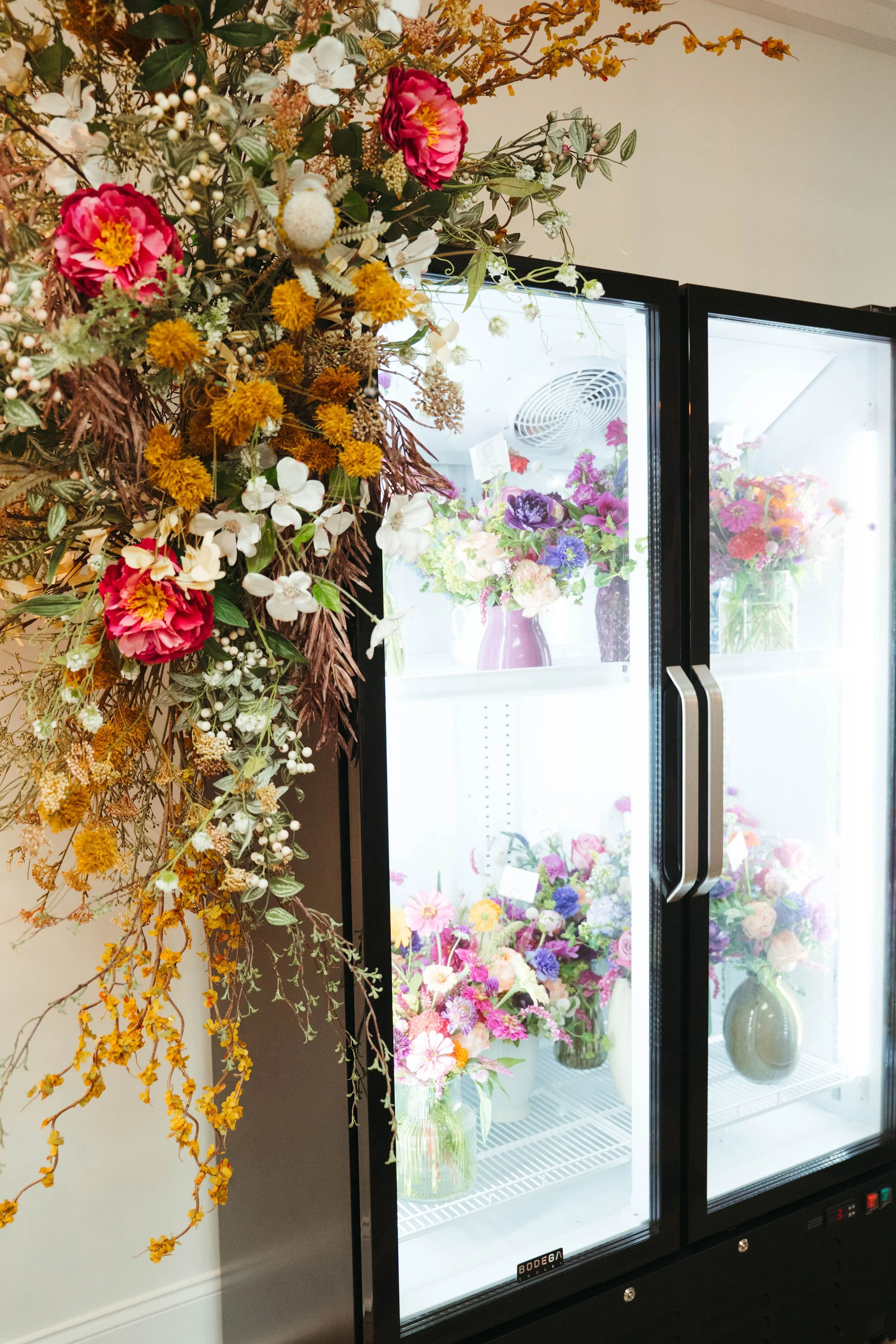 A large floral display with pink, white, yellow, and purple flowers positioned next to a glass-door refrigerator filled with more colorful flowers in vases.