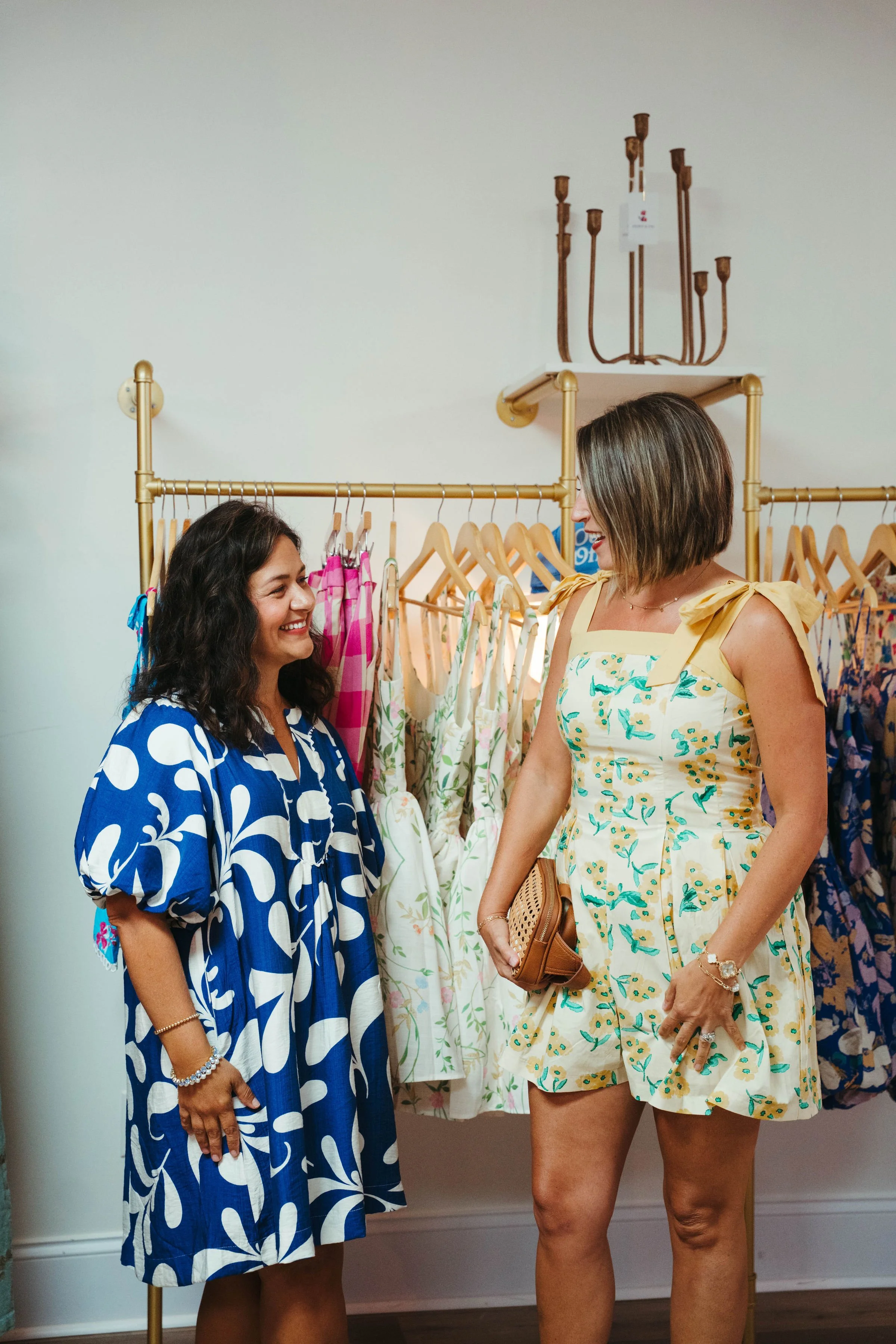 Two women are shopping for summer dresses in a boutique, smiling and talking to each other, surrounded by colorful dresses on a clothing rack.
